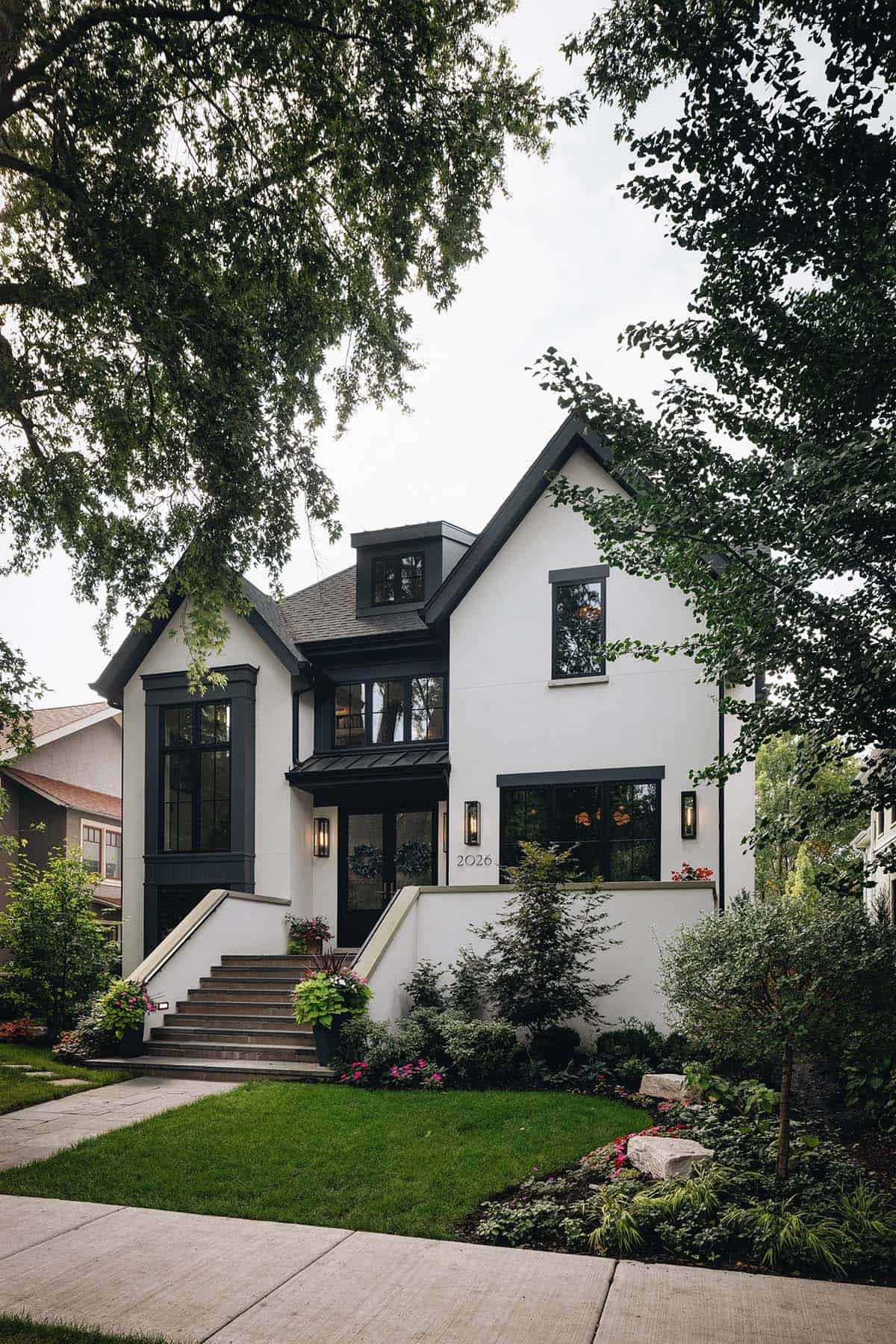 White stucco home exterior with black window trim, steep gabled roofline, stone entry stairs, and lush front garden landscaping
