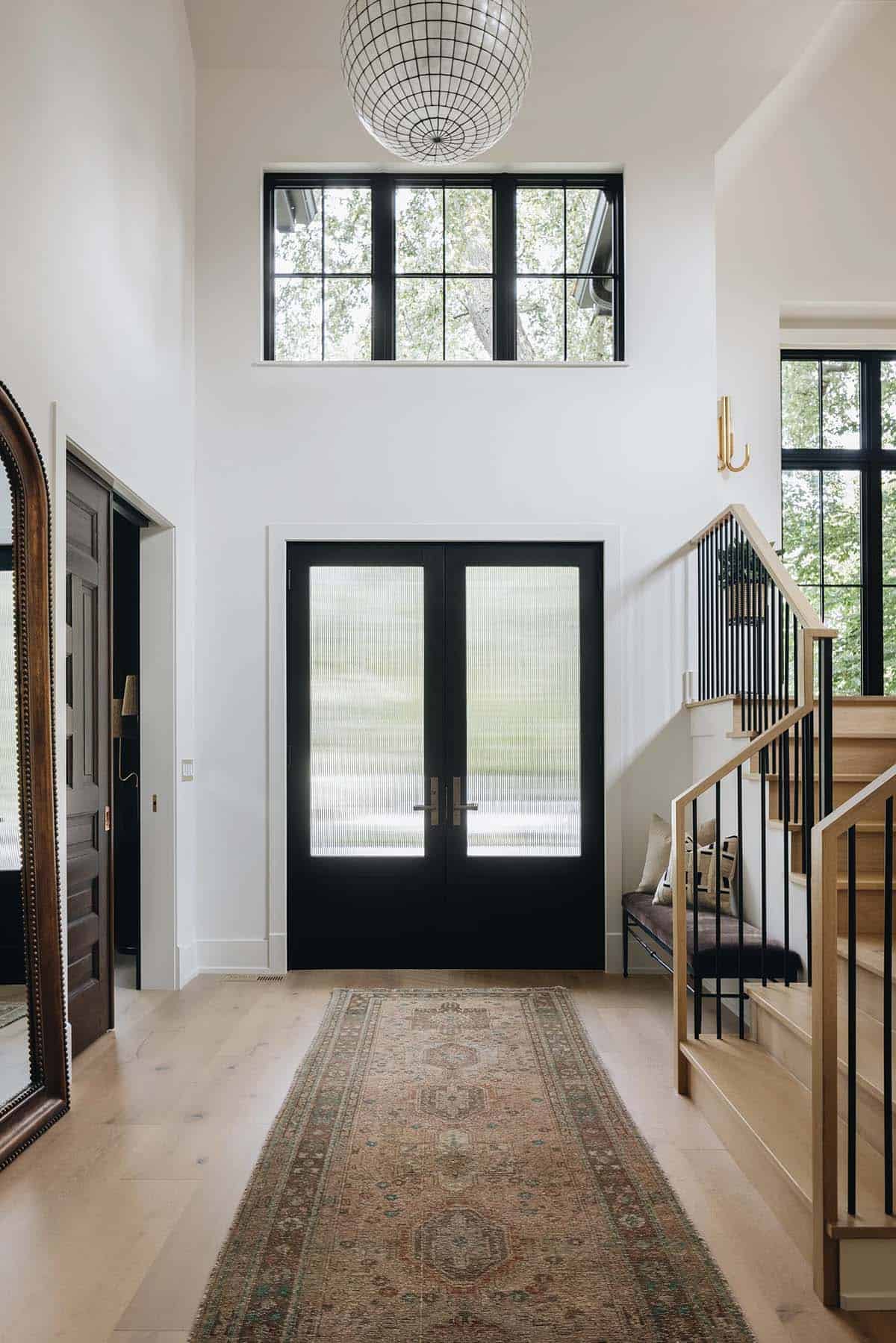 Two-story entry foyer with black French doors, reeded glass, globe pendant light, vintage runner rug, and open staircase with black metal balusters