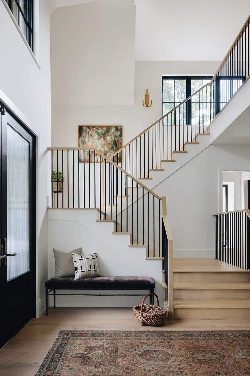 Bright foyer with open-rise staircase, natural oak treads, black steel balusters, velvet bench, and abstract painting on white wall
