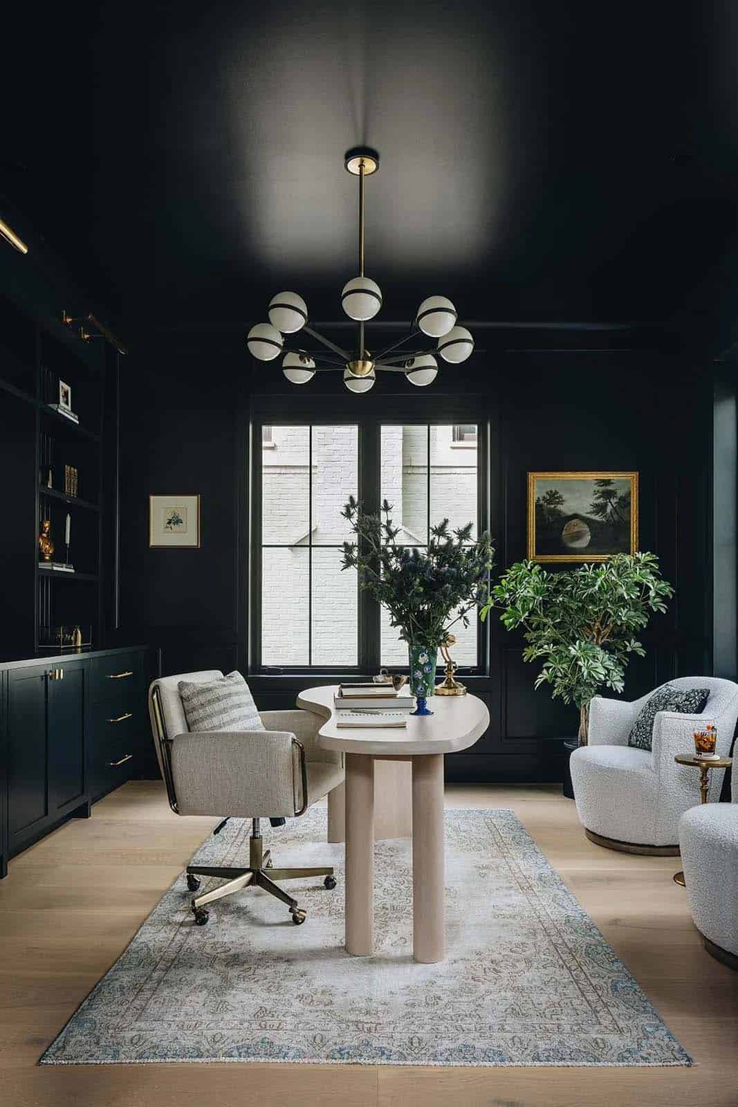 Dramatic dark home office with black painted walls and ceiling, sculptural sputnik chandelier, curved plaster desk, and built-in dark green cabinetry