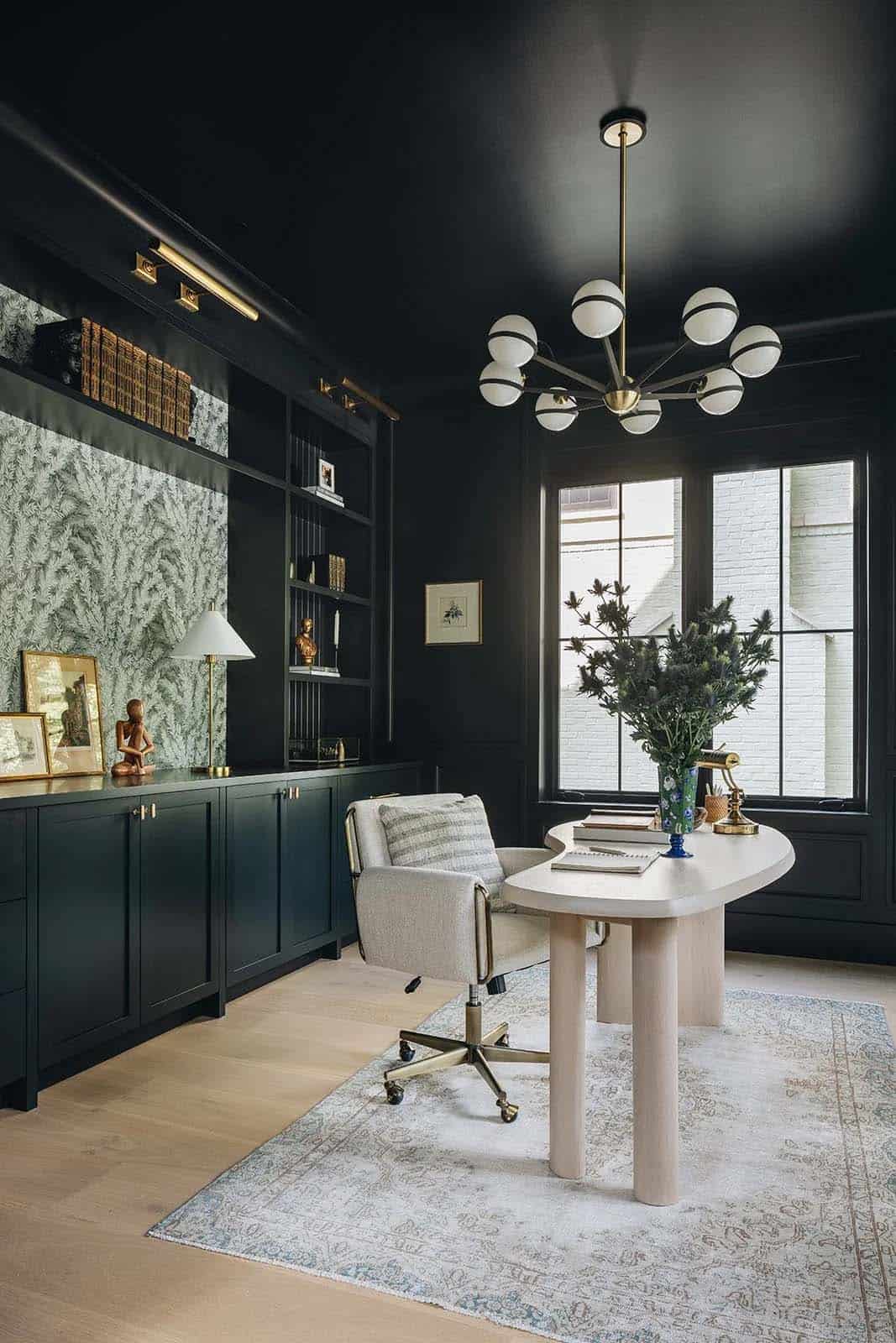 Dark home office with black ceiling, botanical wallpaper, floor-to-ceiling built-ins, and a curved plaster desk topped with a large plant arrangement