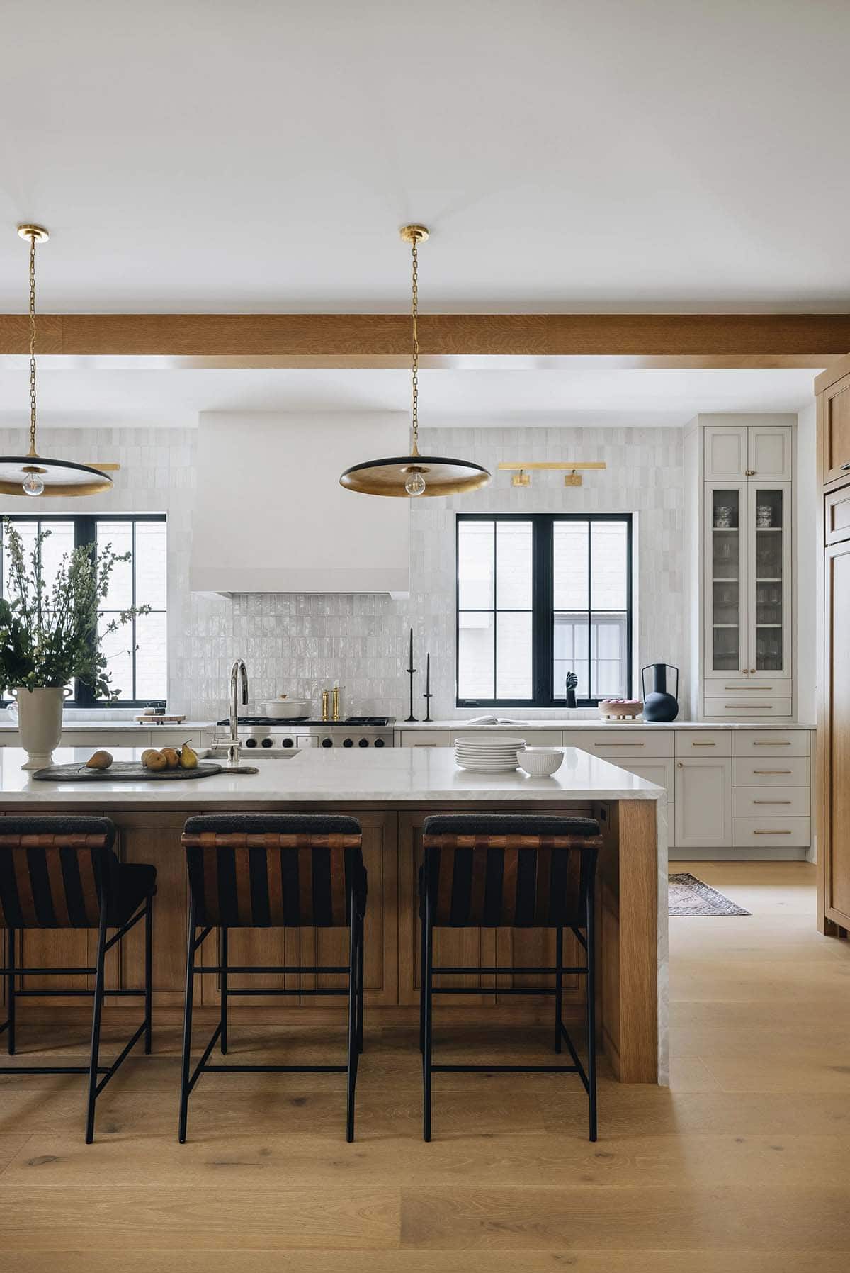 Kitchen island with white marble waterfall countertop, natural oak base, woven leather barstools, brass disc pendants, and exposed ceiling beam
