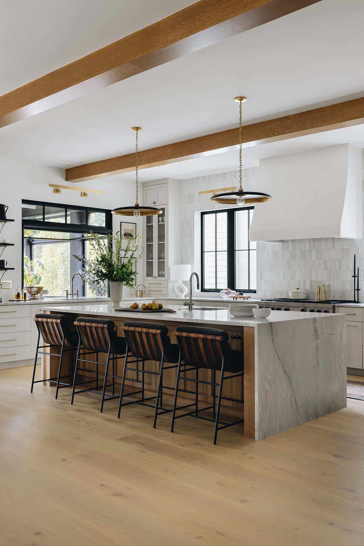 Wide kitchen view with large marble island, natural oak ceiling beams, brass disc pendants, and zellige tile backsplash behind a Wolf range