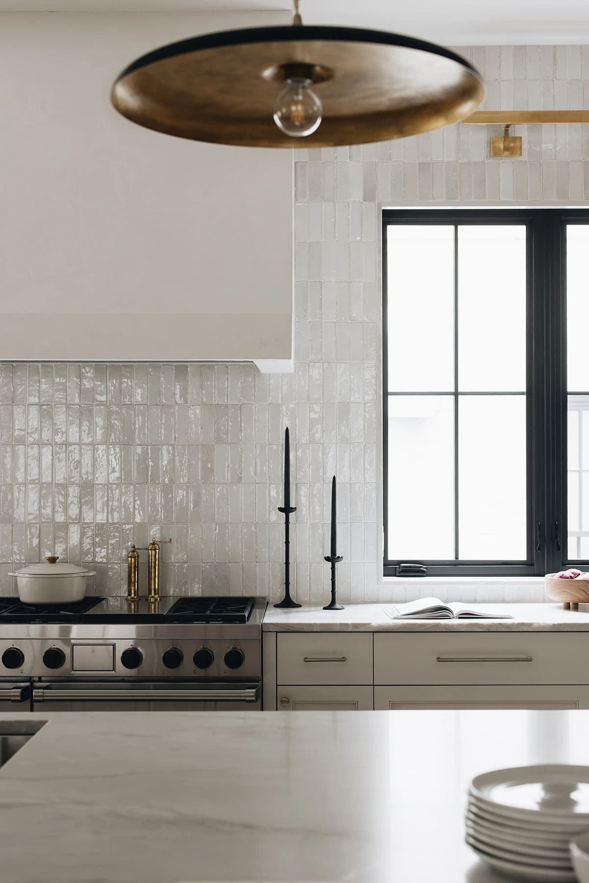 Detail of kitchen cooking wall with zellige tile backsplash, brass faucet, black taper candles, and a brass disc pendant overhead