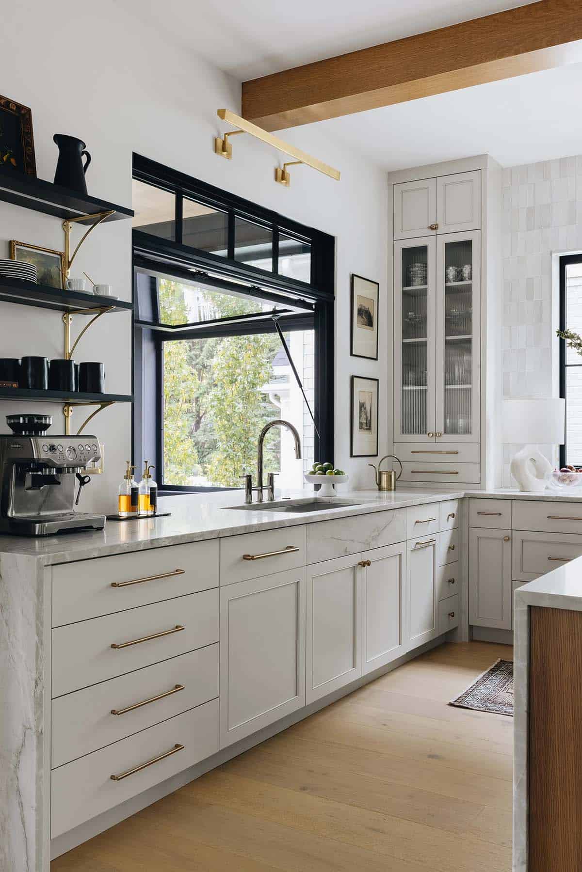 Kitchen sink wall with cream shaker cabinets, brass hardware, black swing-out casement window, open black shelving, and espresso machine on the counter
