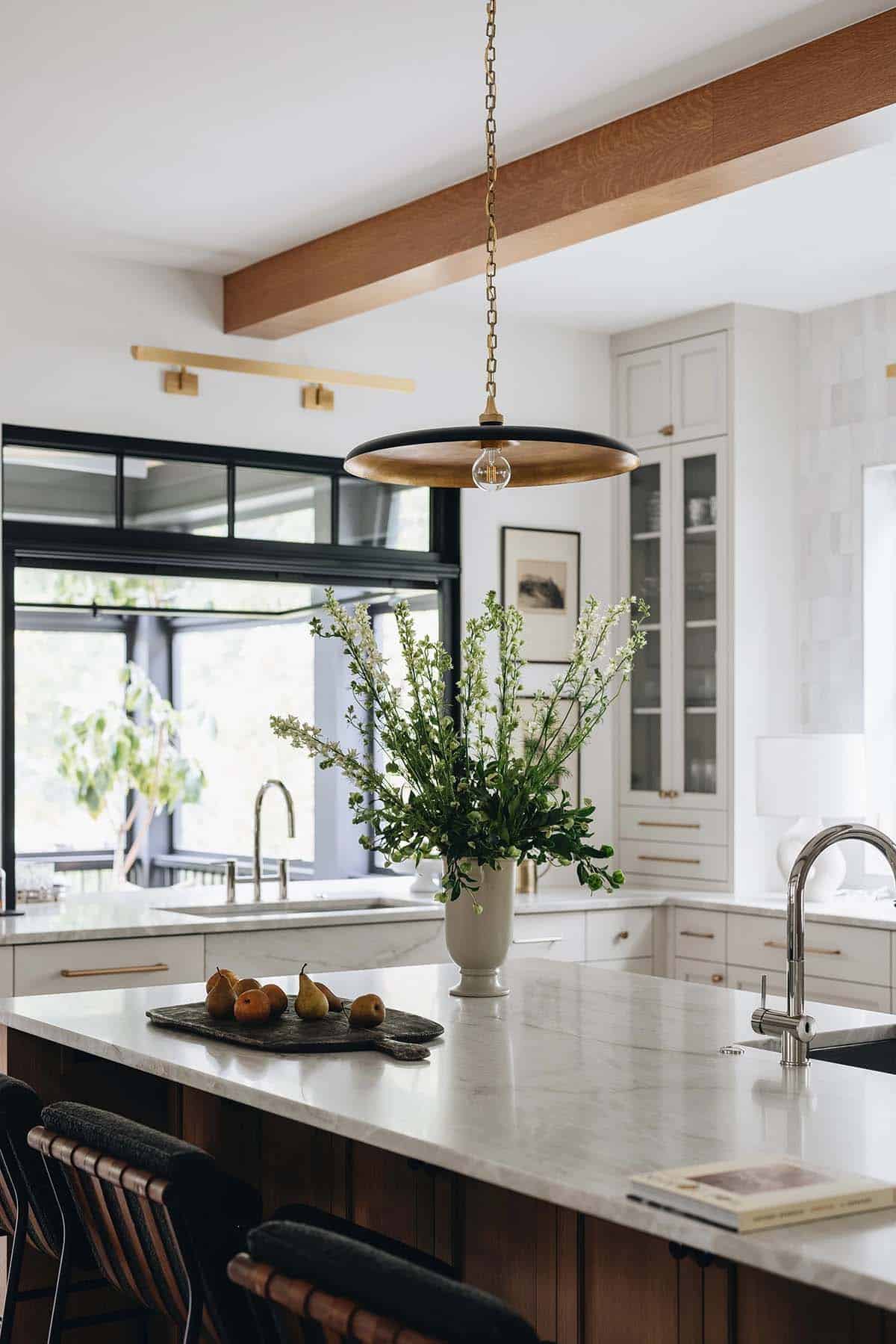 Kitchen island with white marble top, brass disc pendant, and a large floral arrangement in a white ceramic vase beside an open casement window