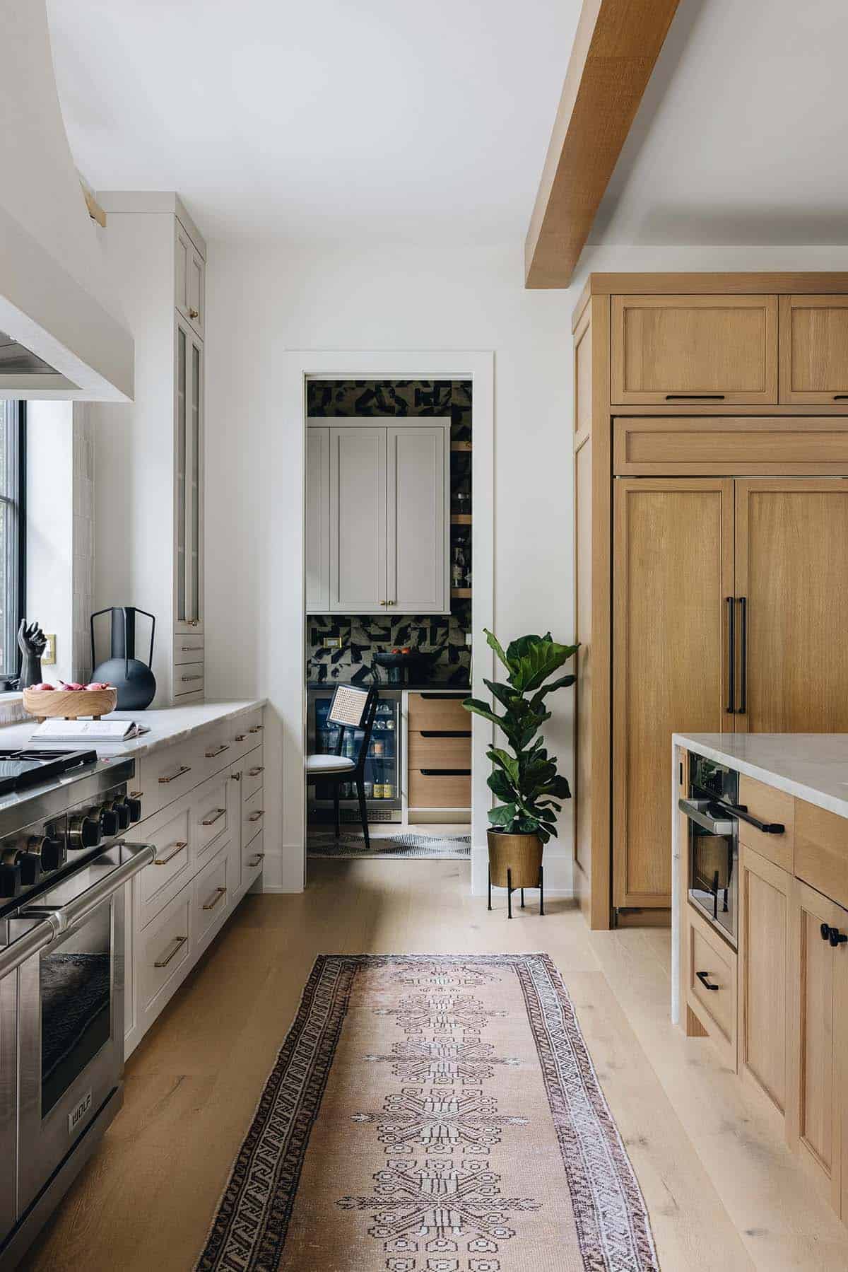 Kitchen view toward pantry doorway with bold graphic wallpaper, cream cabinets, and a fiddle-leaf fig plant beside the oak refrigerator panel