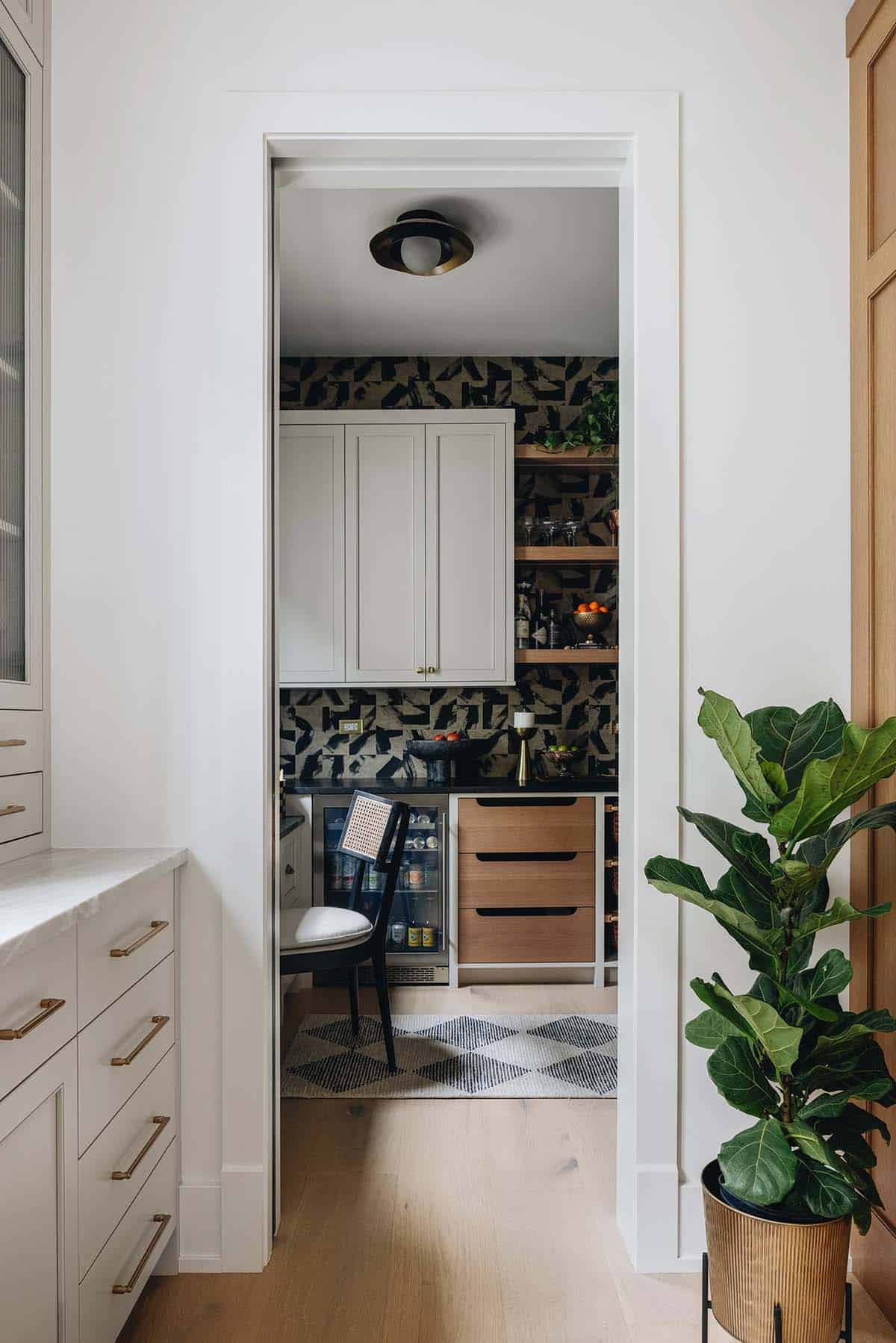 Doorway view into a pantry with dark graphic wallpaper, cream cabinetry, open wood shelving, and a beverage fridge below the counter