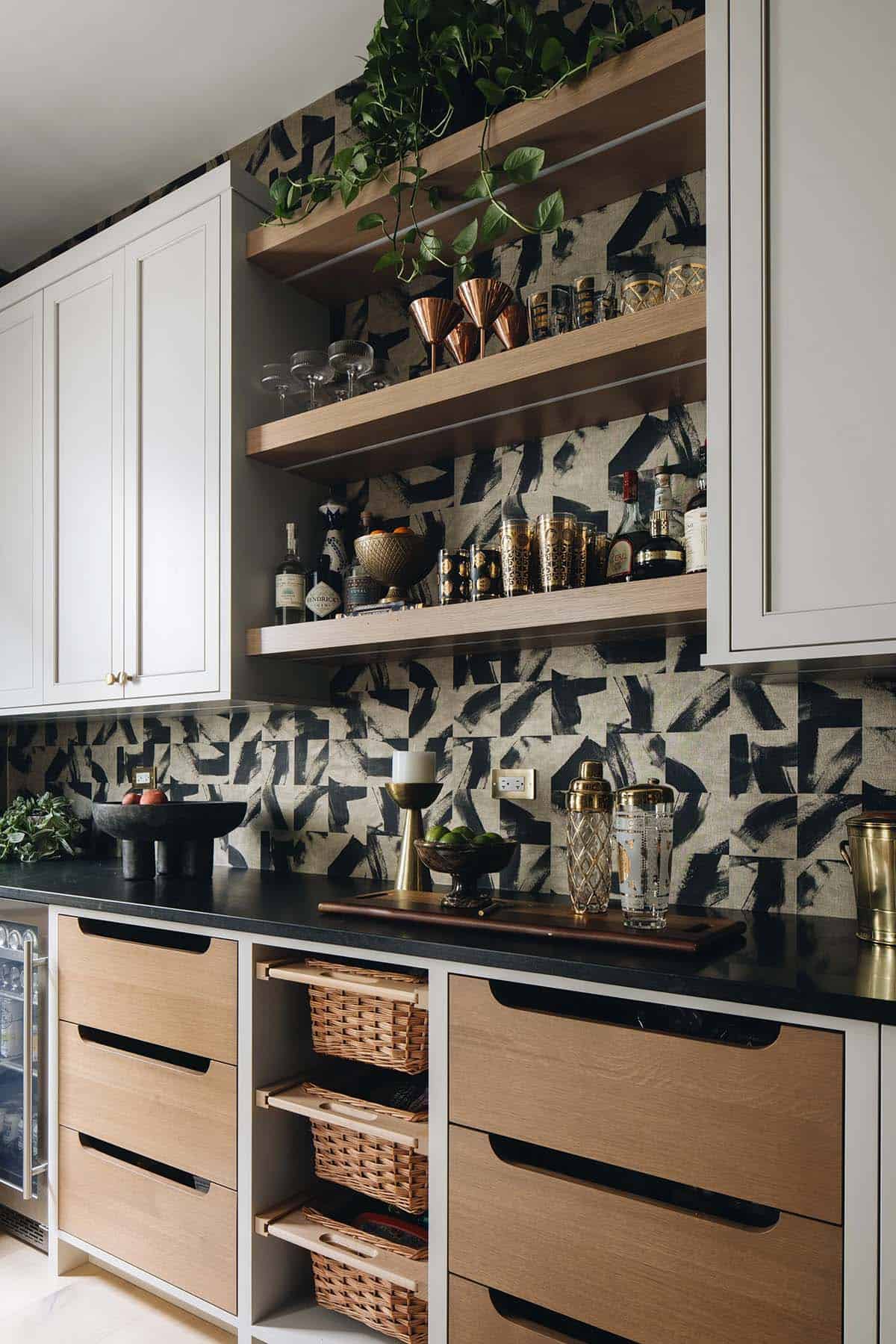 Butler's pantry bar wall with bold graphic wallpaper, open oak shelving stocked with barware and spirits, black granite countertop, and woven basket storage below