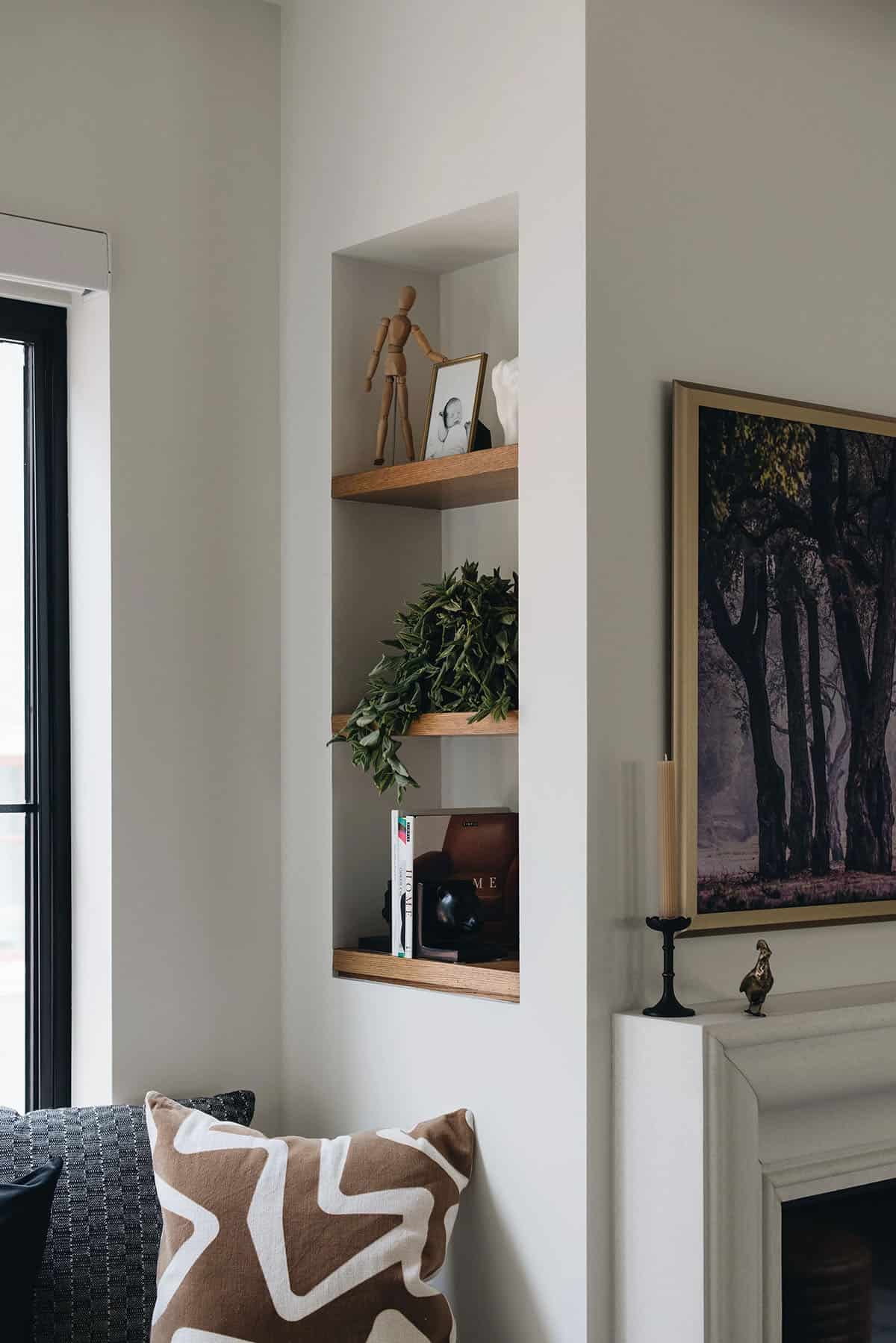 Built-in wall niche with three oak shelves styled with a wooden figure, trailing greenery, framed photo, and stacked books beside the fireplace
