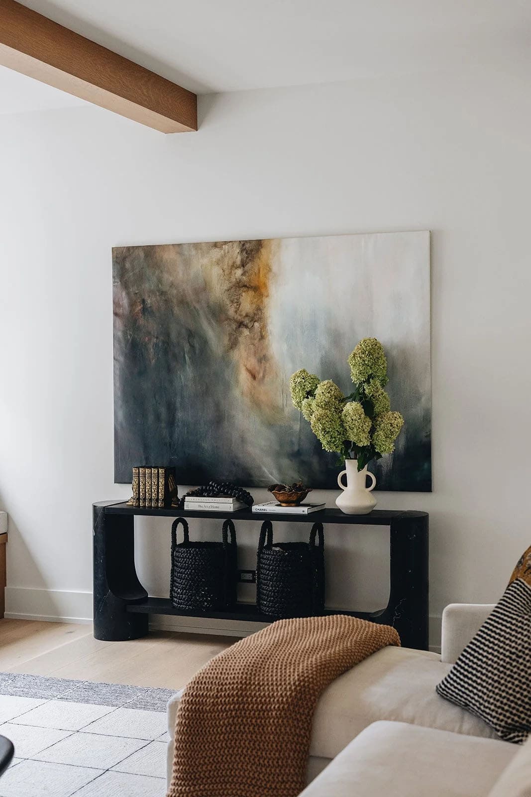Living room vignette with a large abstract painting in blue and gold tones above a sculptural black console table with hydrangea arrangement