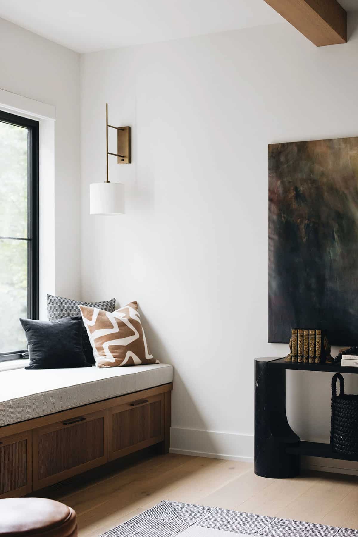 Corner of living room with an oak window seat, dark velvet and graphic print throw pillows, a brass wall sconce, and a partial view of the console table