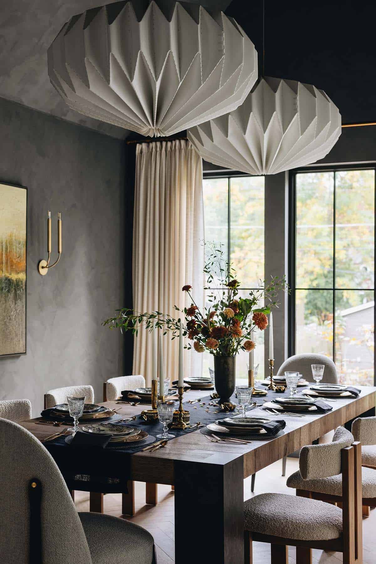 Dining room table set with a dahlia centerpiece in a bronze vase, white taper candles, crystal stemware, and dark linen napkins under two origami pendants