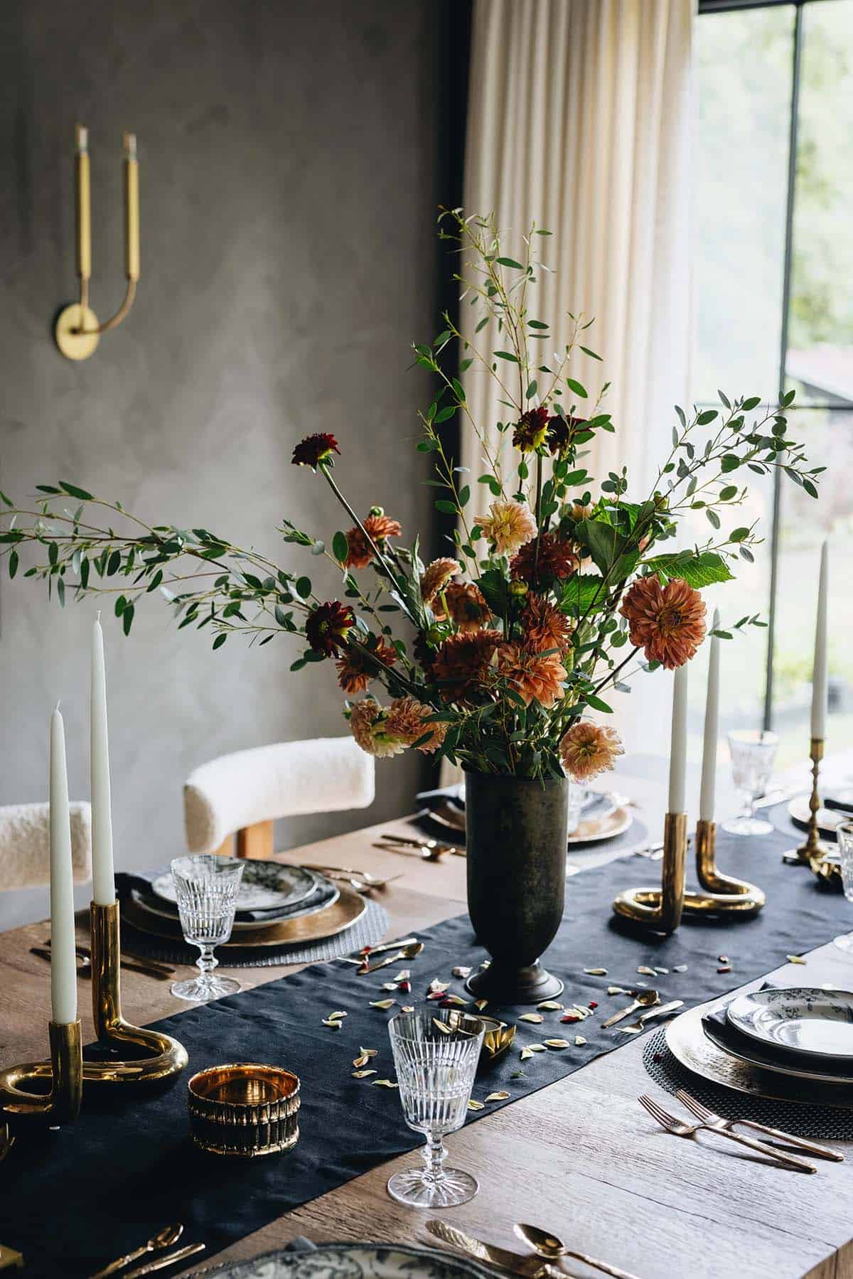 Dining room table detail with a tall dahlia and eucalyptus centerpiece, white taper candles in brass holders, layered place settings, and crystal goblets