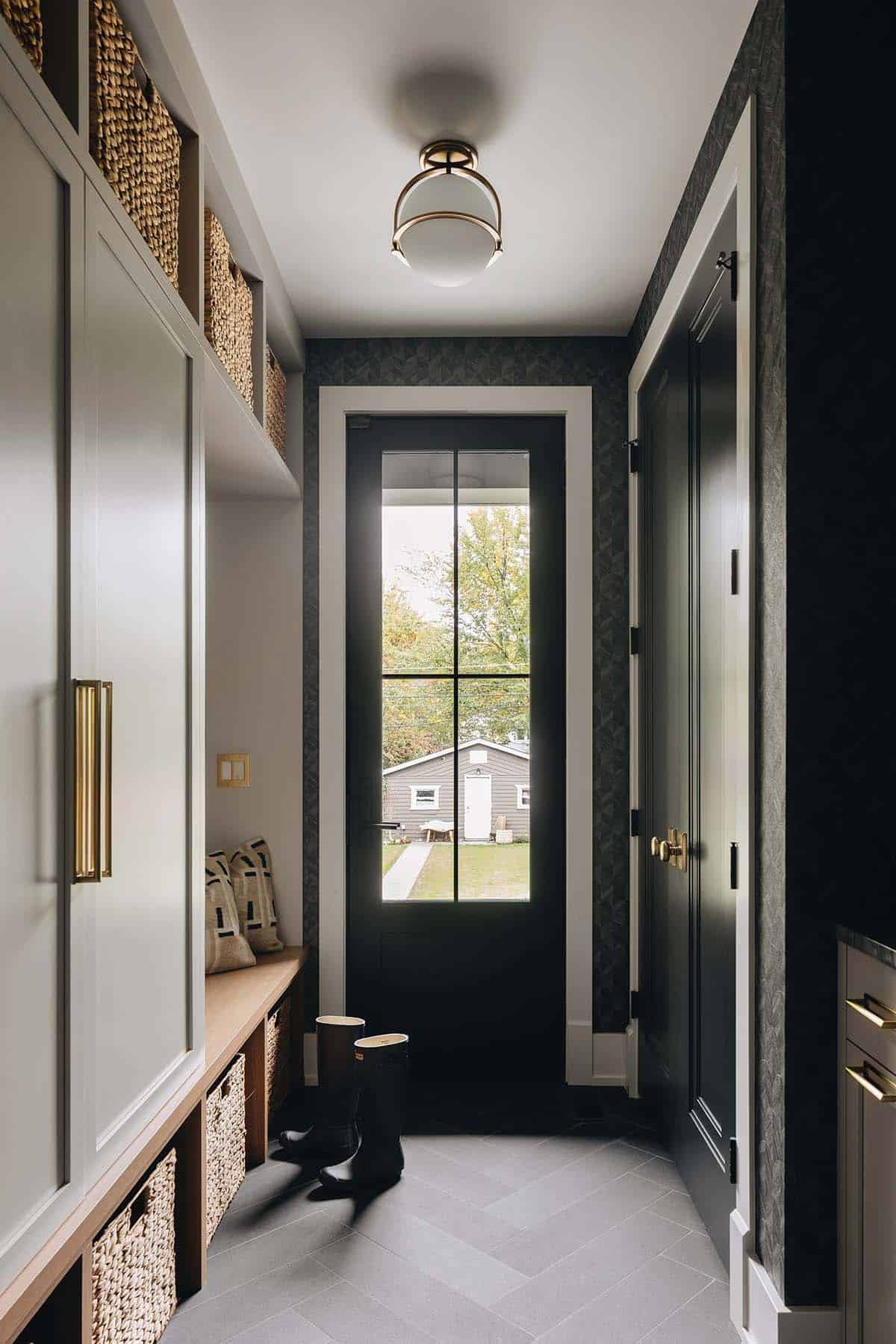 Mudroom hallway with herringbone tile floor, cream cabinetry, dark wallpaper, glazed black back door, and a brass-accented ceiling light