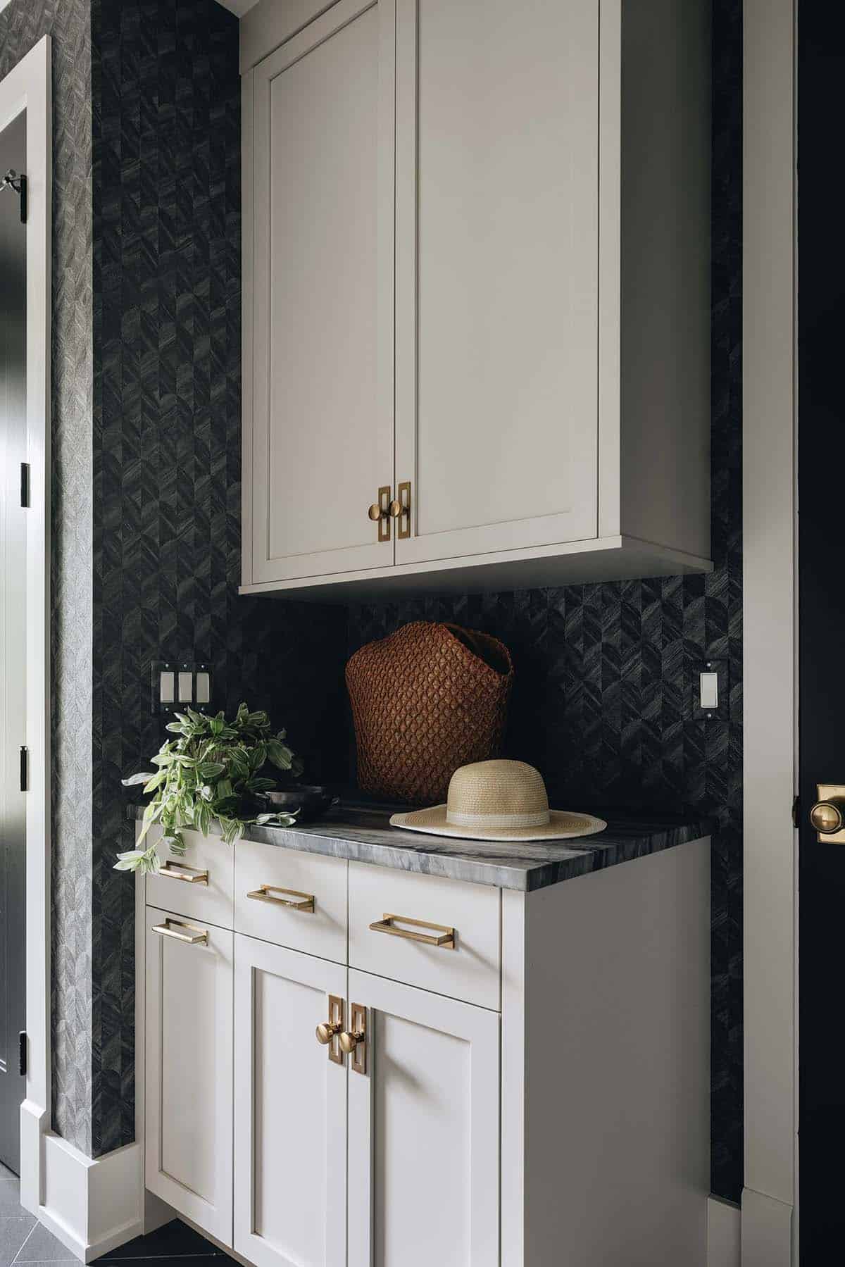 Mudroom detail with cream upper and lower cabinetry, dark geometric wallpaper, marble countertop, brass hardware, and a straw hat and woven bag on the counter