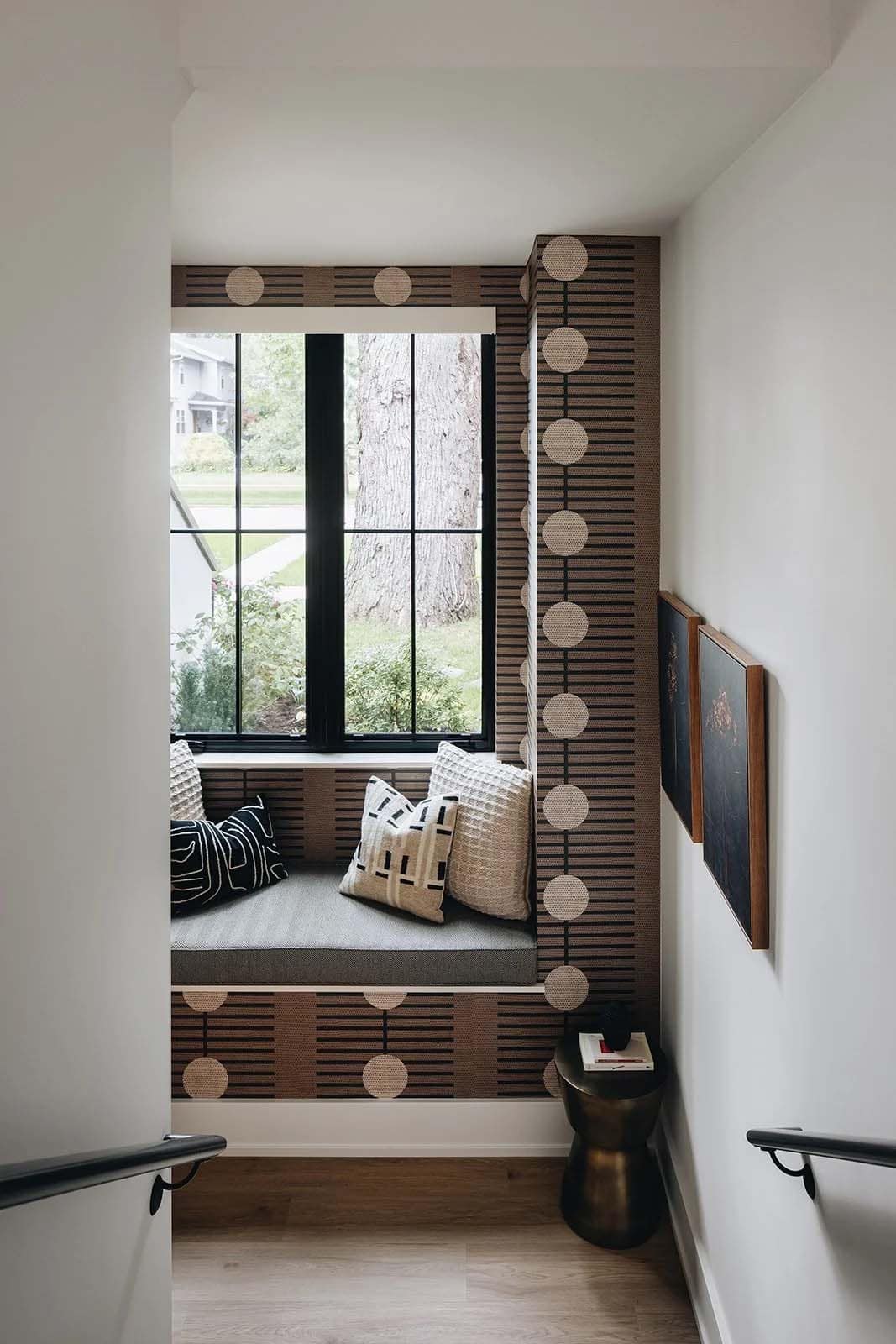 Staircase landing reading nook with a bold geometric brown and tan wallpaper, built-in window seat with graphic pillows, and a brass side table