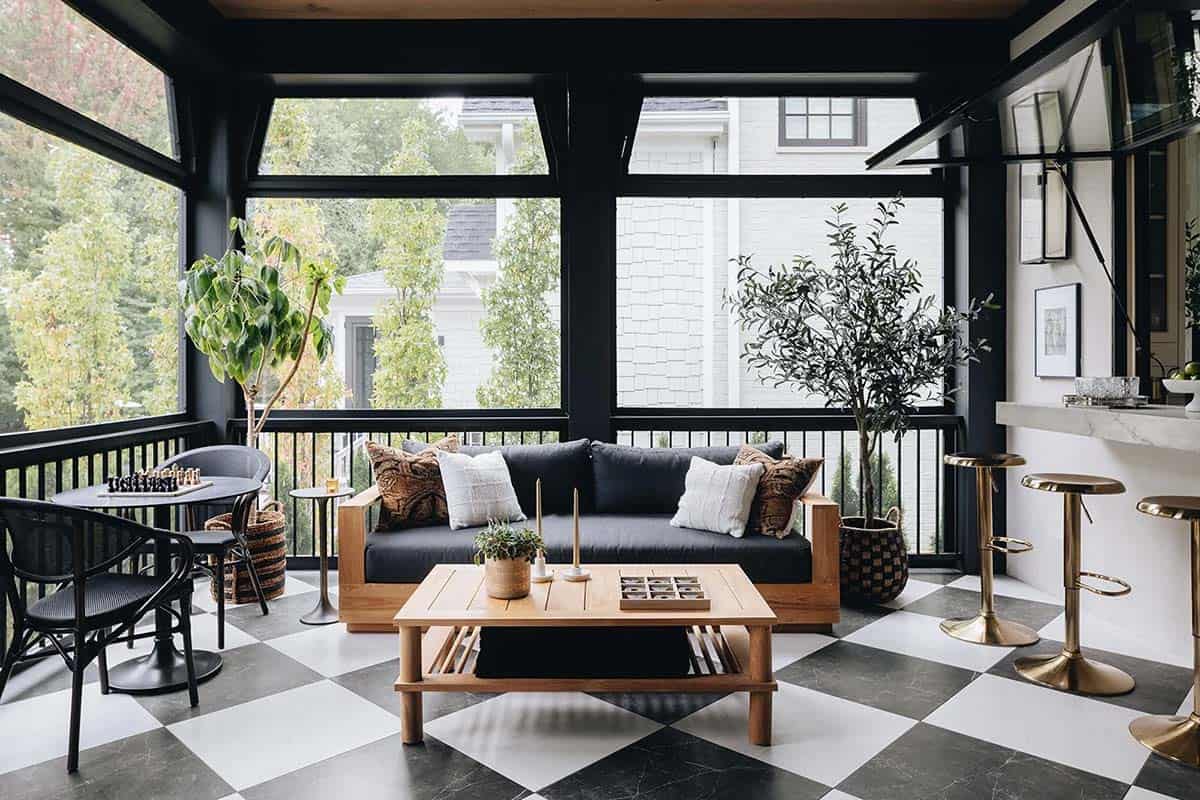 Screened porch with black-framed panels, checkerboard tile floor, teak sofa and coffee table, potted olive tree, and brass bar stools at a marble pass-through counter