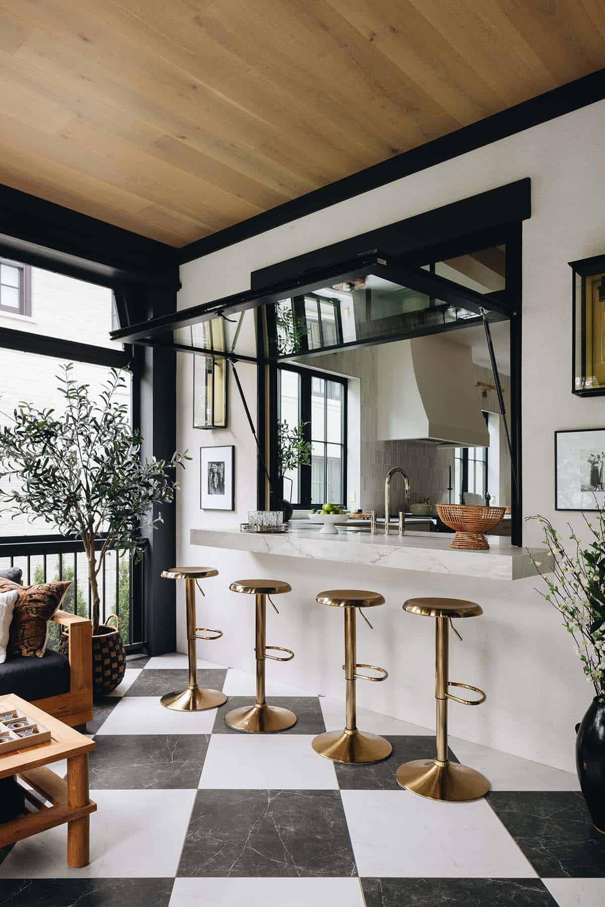 Screened porch pass-through bar with a lift-up glass window, marble countertop, brass bar stools, oak ceiling planks, and a view into the kitchen