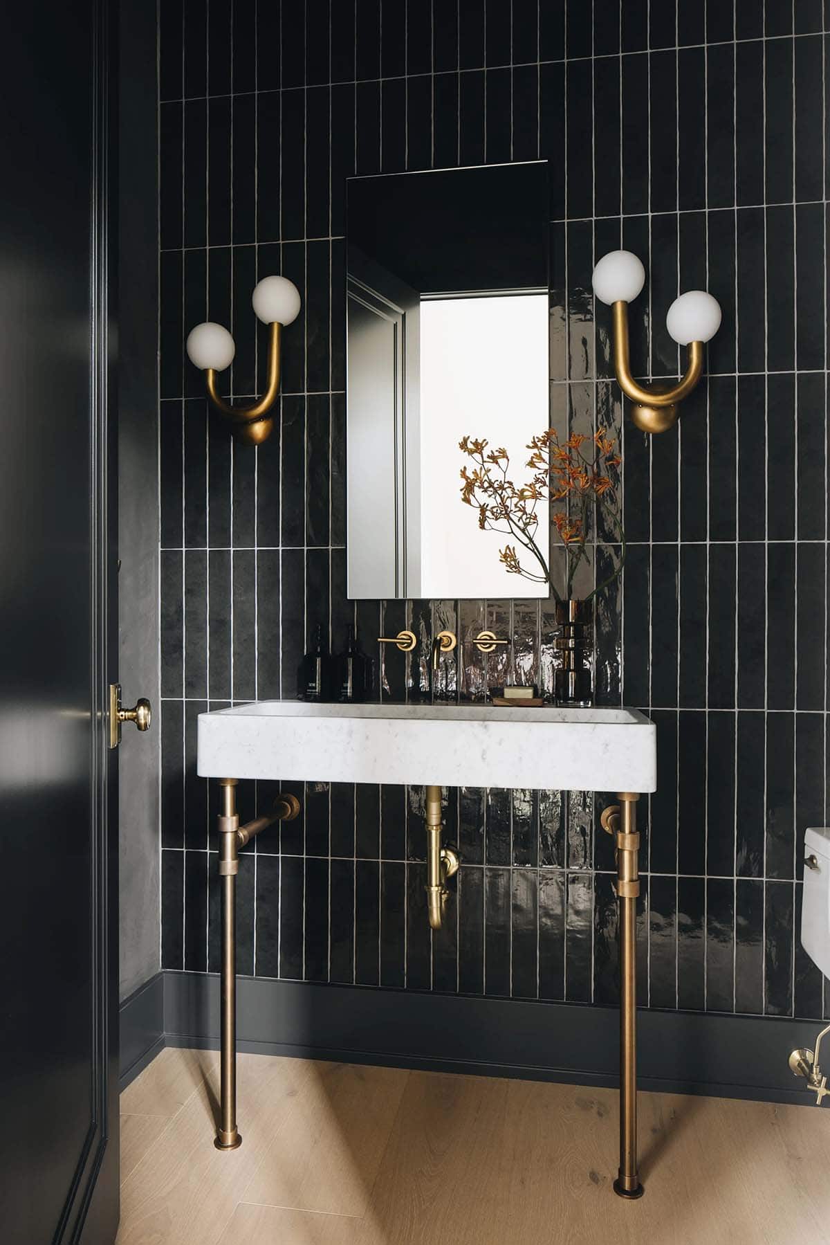 Powder bath with floor-to-ceiling black vertical tile, marble console sink on brass legs, globe sconces, and a recessed medicine cabinet mirror