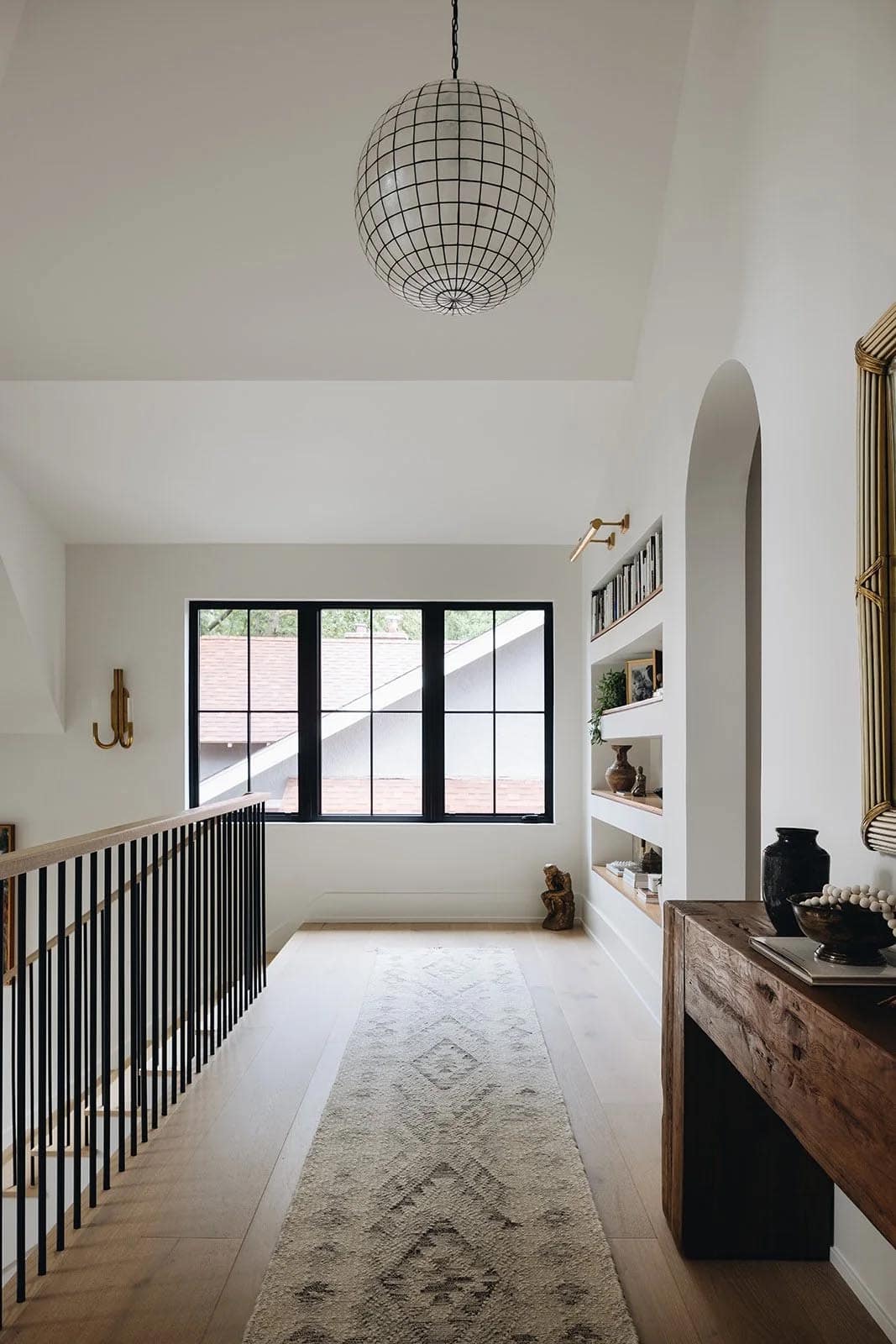 Upper hallway with a grid-pattern globe pendant, arched built-in bookshelf niche, runner rug, black steel stair railing, and a rustic wood console table