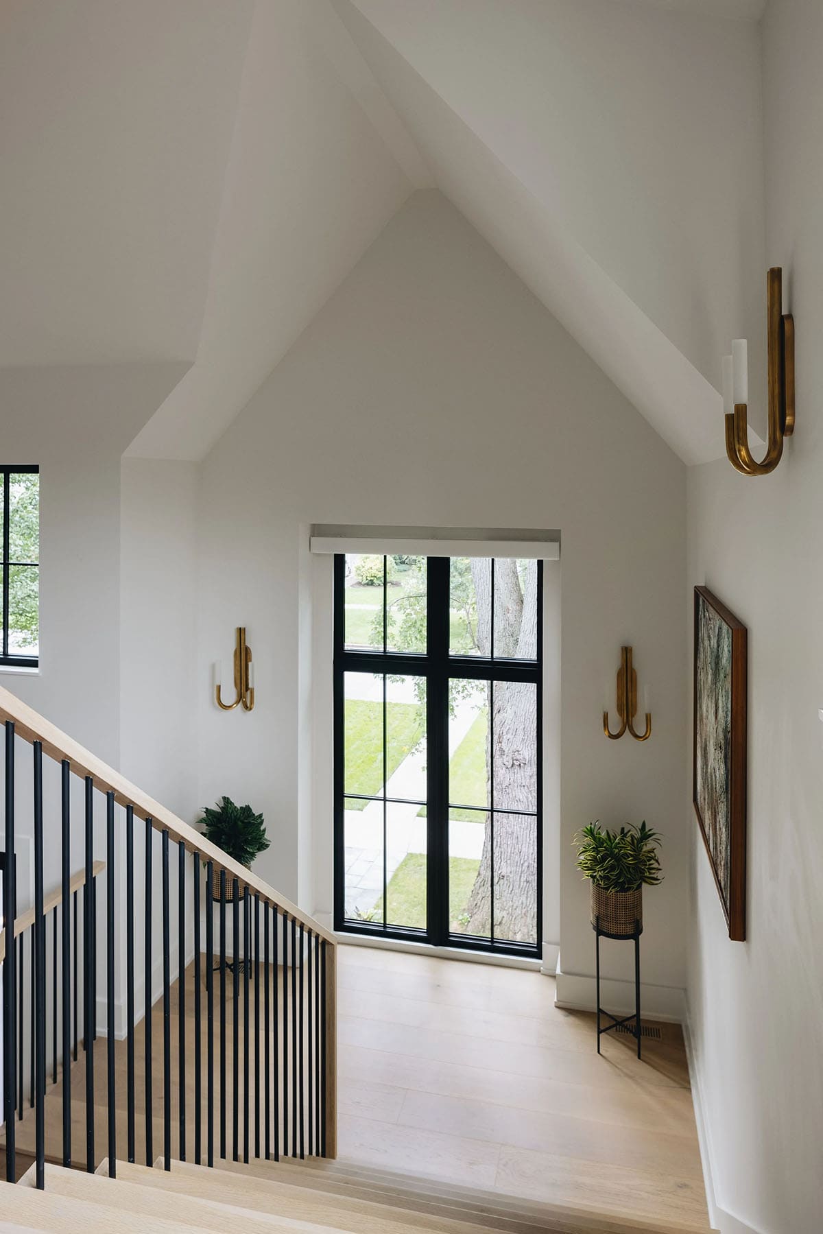 Staircase landing with vaulted white ceiling, tall black-framed grid window, brass hook sconces, and a potted plant on a black metal stand