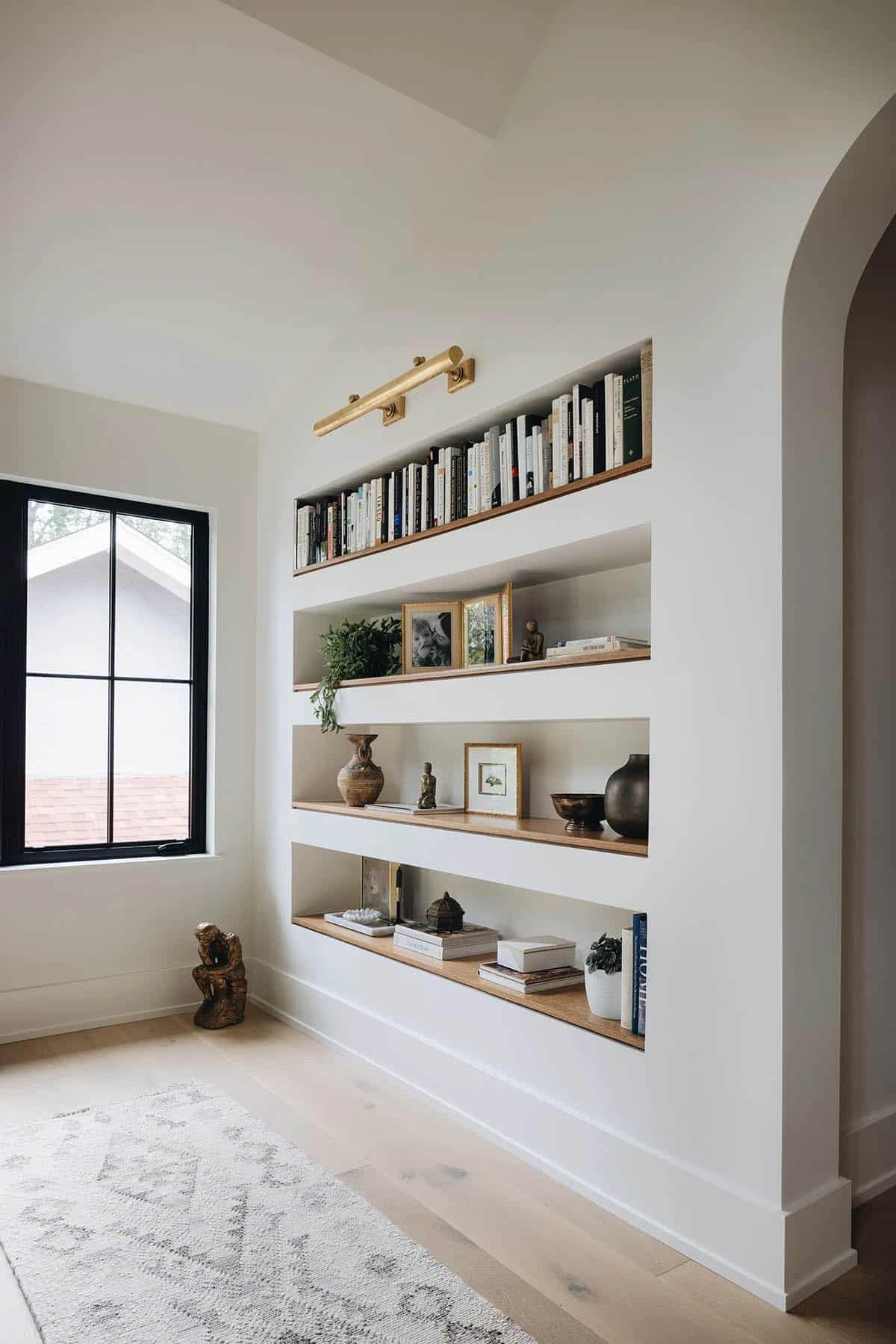 Hallway built-in bookcase niche with four oak shelves styled with books, framed photos, ceramics, and vases, lit by a brass picture light