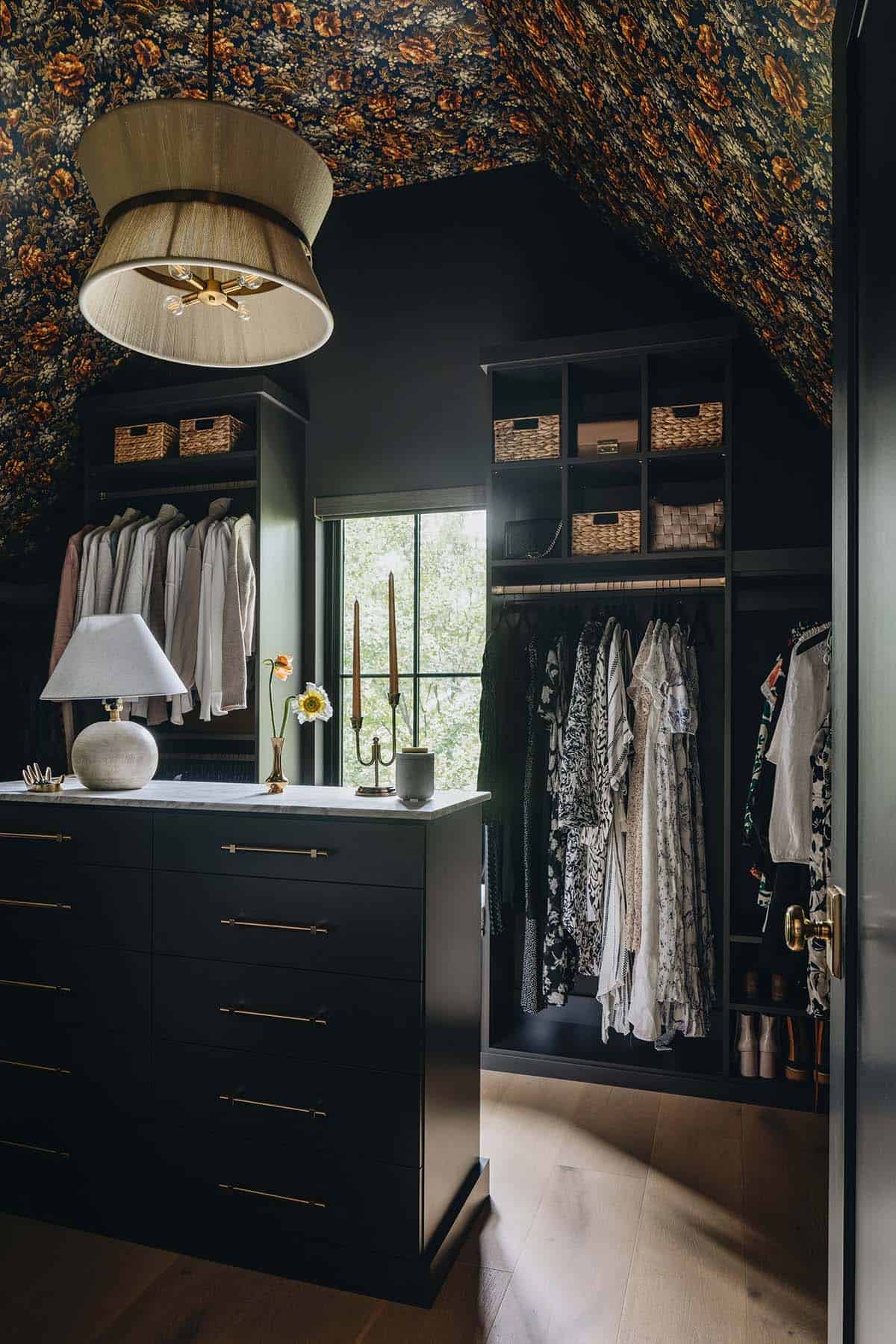 Walk-in closet with dark navy walls, bold floral wallpapered ceiling, black built-in cabinetry with brass hardware, a marble-topped island dresser, and hanging clothes