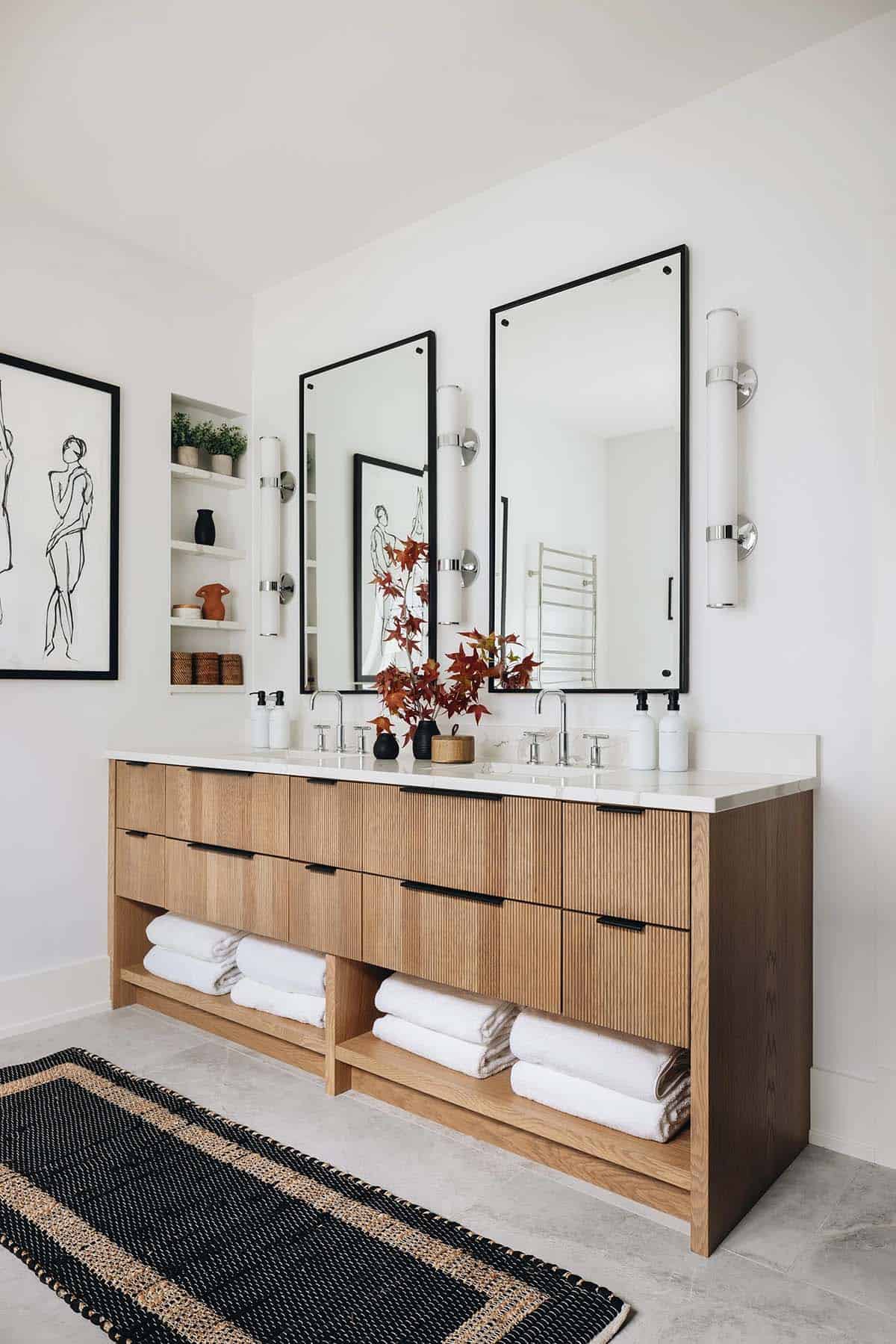 Primary bathroom double vanity with fluted oak cabinet, white marble countertop, dual black-framed mirrors, chrome sconces, and red Japanese maple branch arrangement
