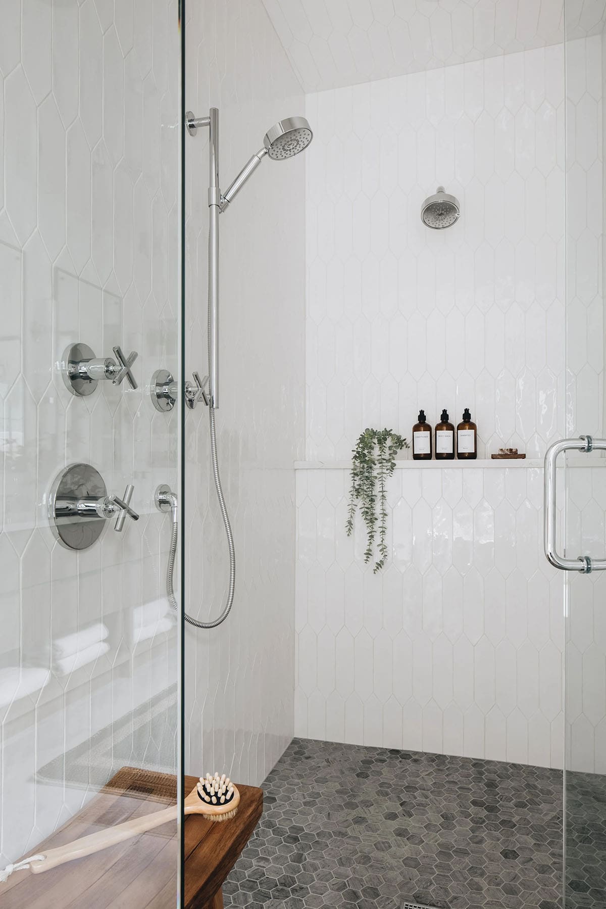 Walk-in shower interior with white picket tile walls, chrome cross-handle fixtures, handheld shower wand, gray hex mosaic floor, and a built-in shelf with amber bath bottles