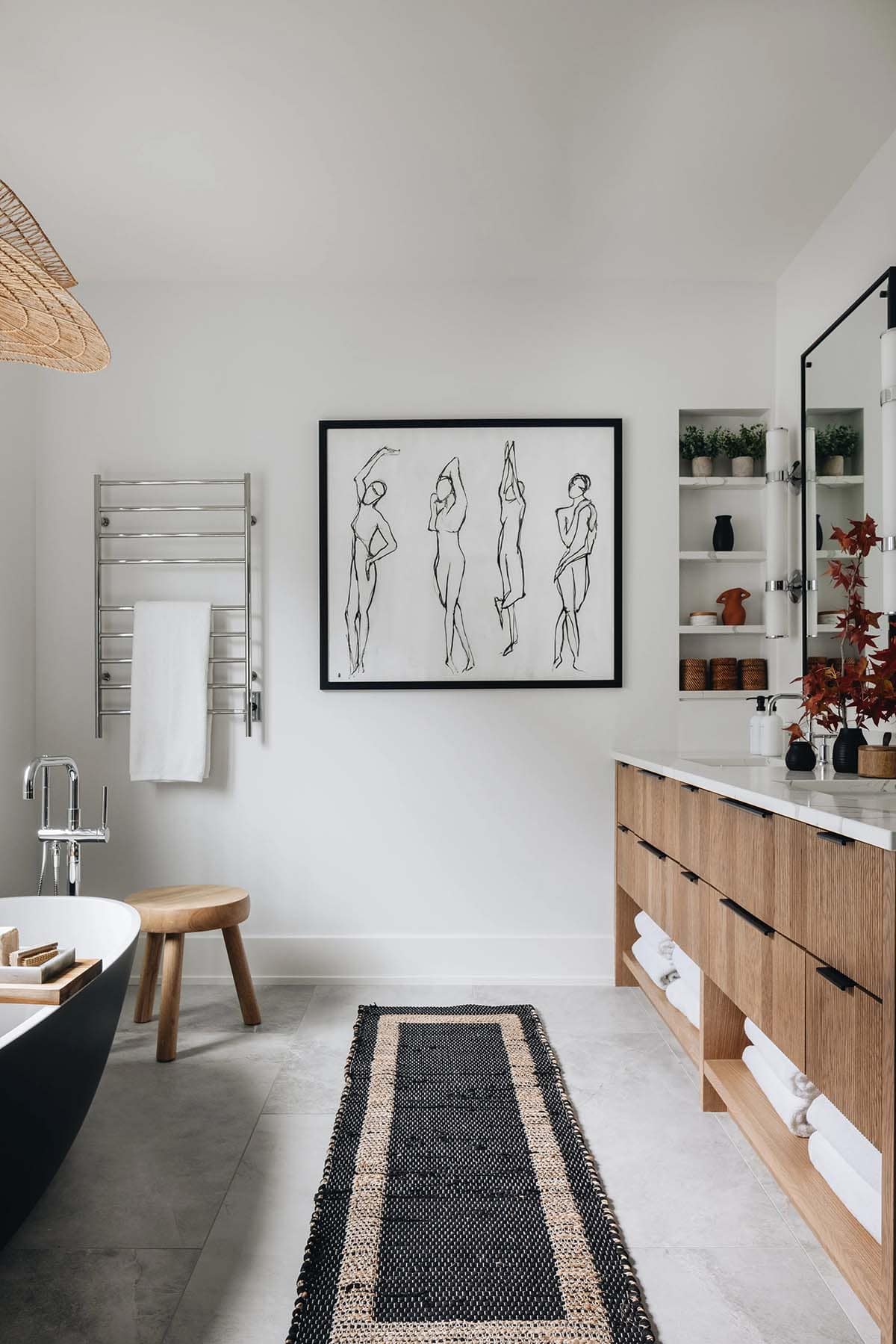 Primary bathroom overview with matte black freestanding tub, chrome towel warmer, line-drawing figure artwork, fluted oak double vanity, and a woven rattan pendant