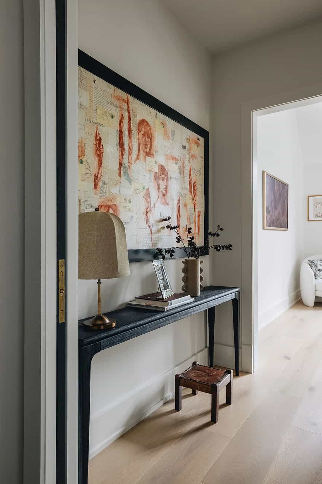 Upper hallway vignette with a large figurative collage artwork, dark console table, dome table lamp, and a view through the doorway into a bedroom