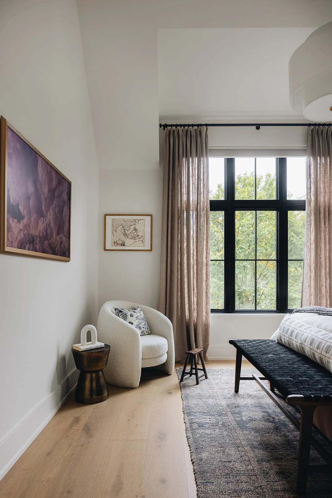 Primary bedroom sitting corner with a boucle swivel chair, brass side table, linen drapes, black-framed grid window, and a landscape painting on the wall