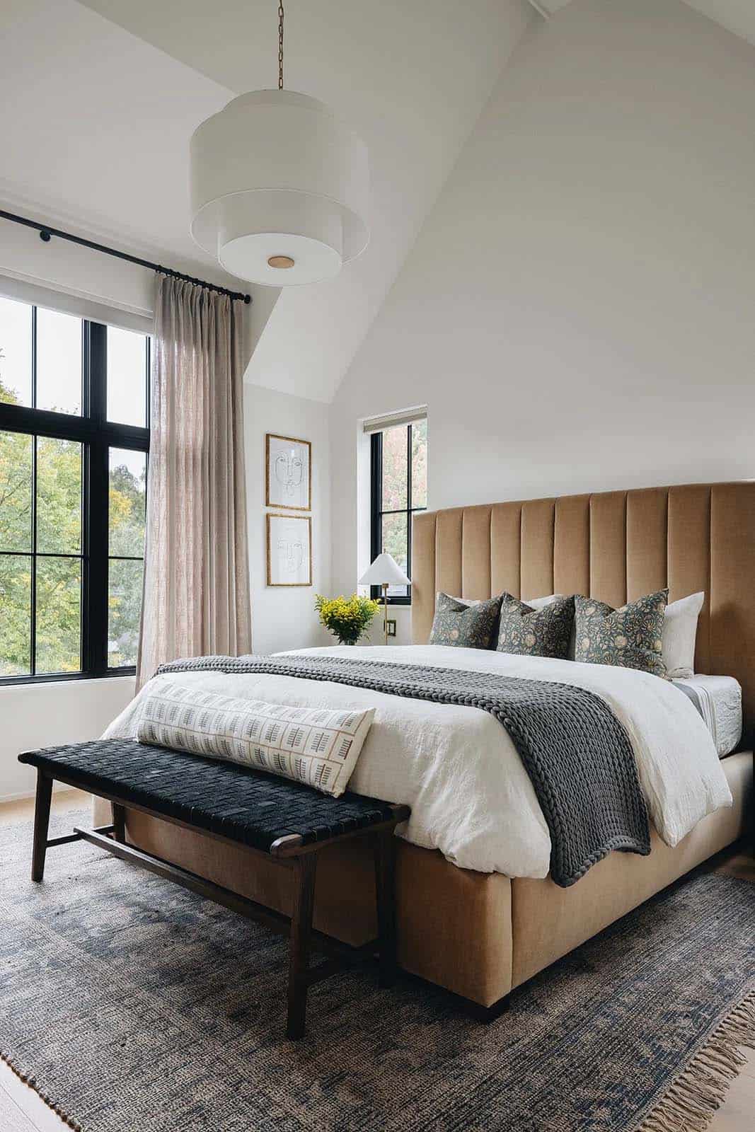 Primary bedroom with a tall caramel velvet channel-tufted headboard, white drum pendant, knit throw, floral print pillows, and black-framed windows overlooking trees