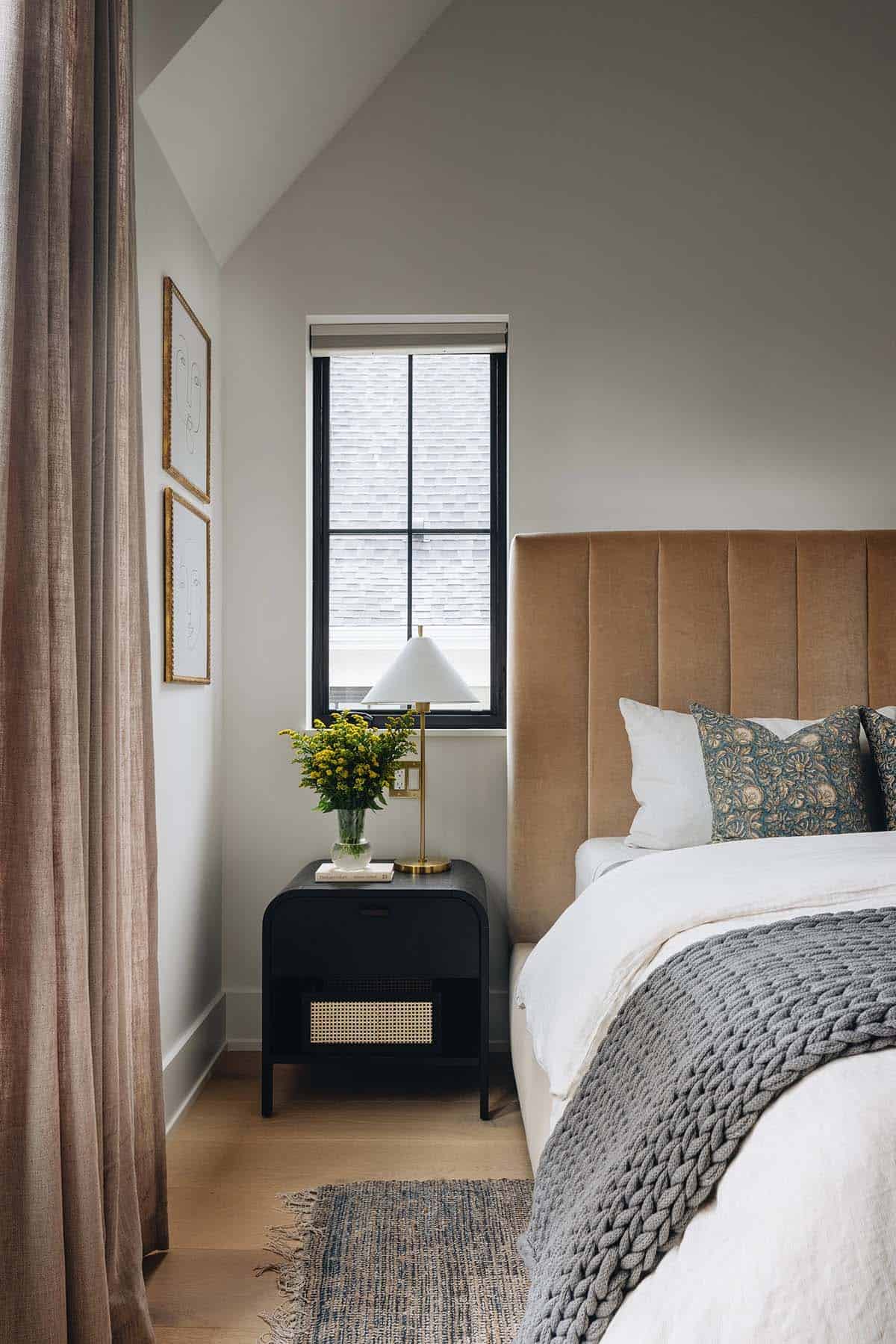 Primary bedroom nightstand detail with a black cane-front bedside table, brass table lamp, yellow wildflower arrangement, and a tall channel-tufted velvet headboard