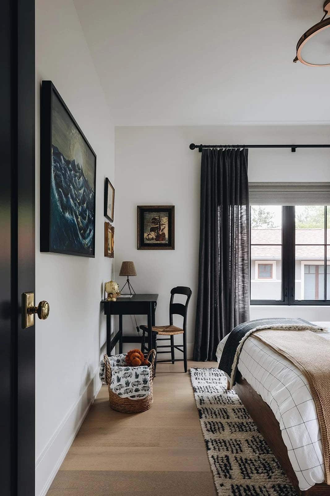 Boy's bedroom with a dark stormy-sea painting gallery wall, a small black writing desk, rush-seat chair, globe lamp, and charcoal linen curtains at a black-framed window