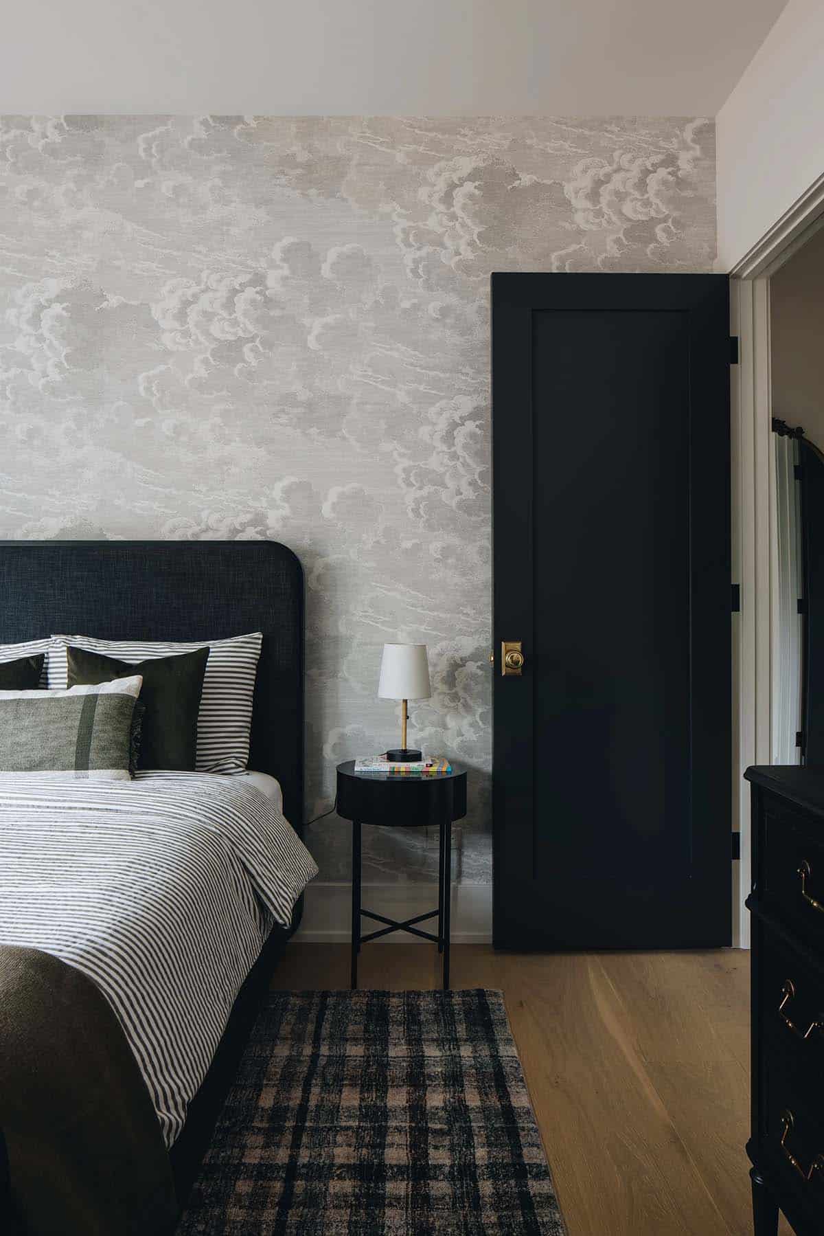Child's bedroom with a cloud-print wallpaper accent wall, dark upholstered headboard, ticking-stripe bedding, black door with brass hardware, and a plaid area rug