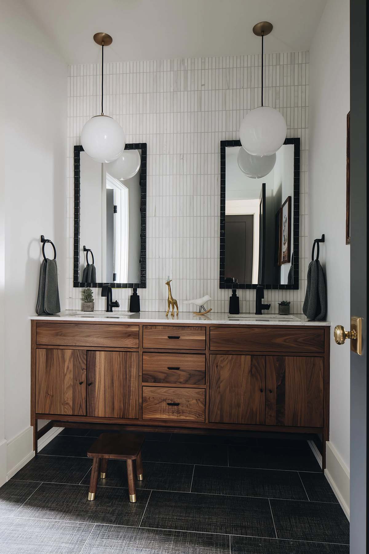 Kids' shared bathroom with a walnut double vanity, white vertical tile backsplash, two black-framed mirrors, globe pendant lights, and dark hexagon floor tile