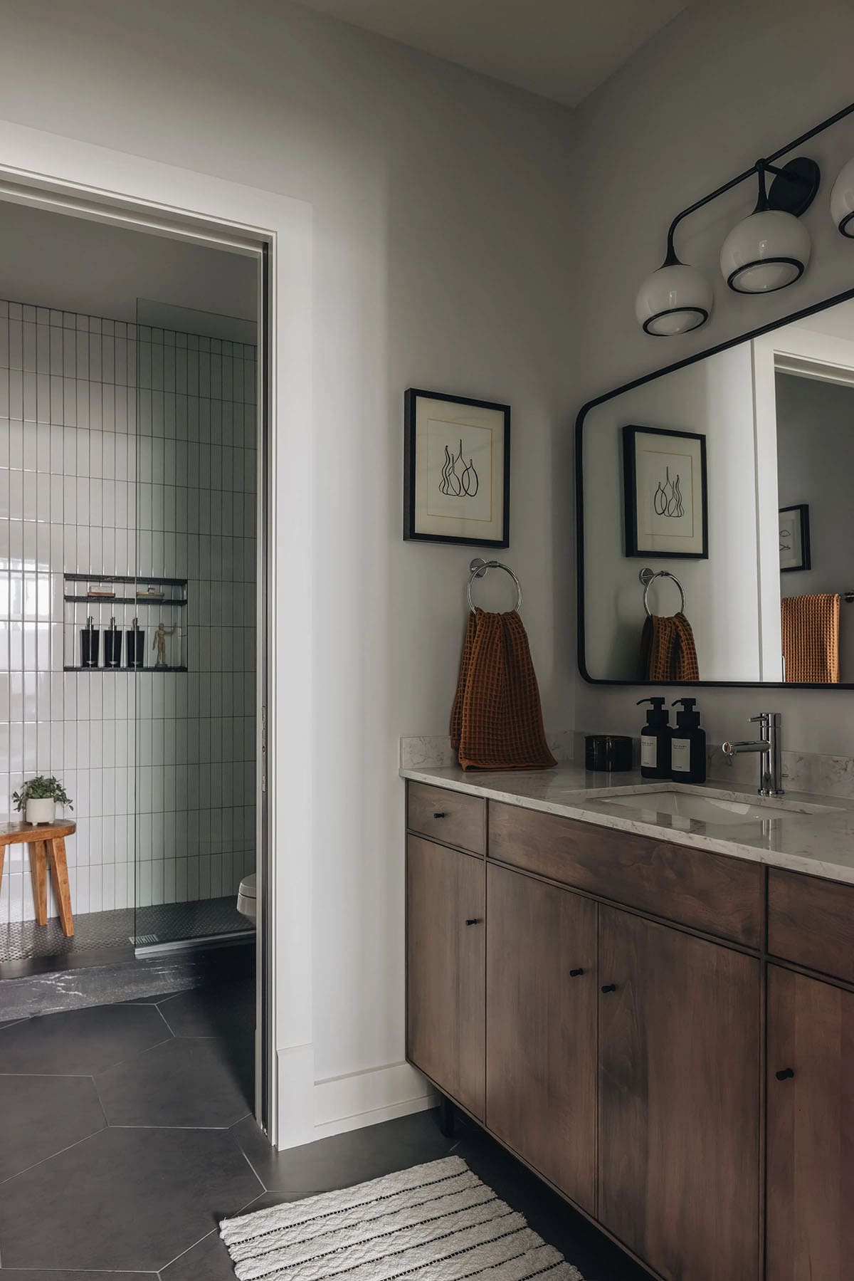 Guest bathroom with a walnut vanity, marble countertop, large black-framed mirror, globe vanity light, and a doorway view into a white-tiled walk-in shower with dark hex floor tile