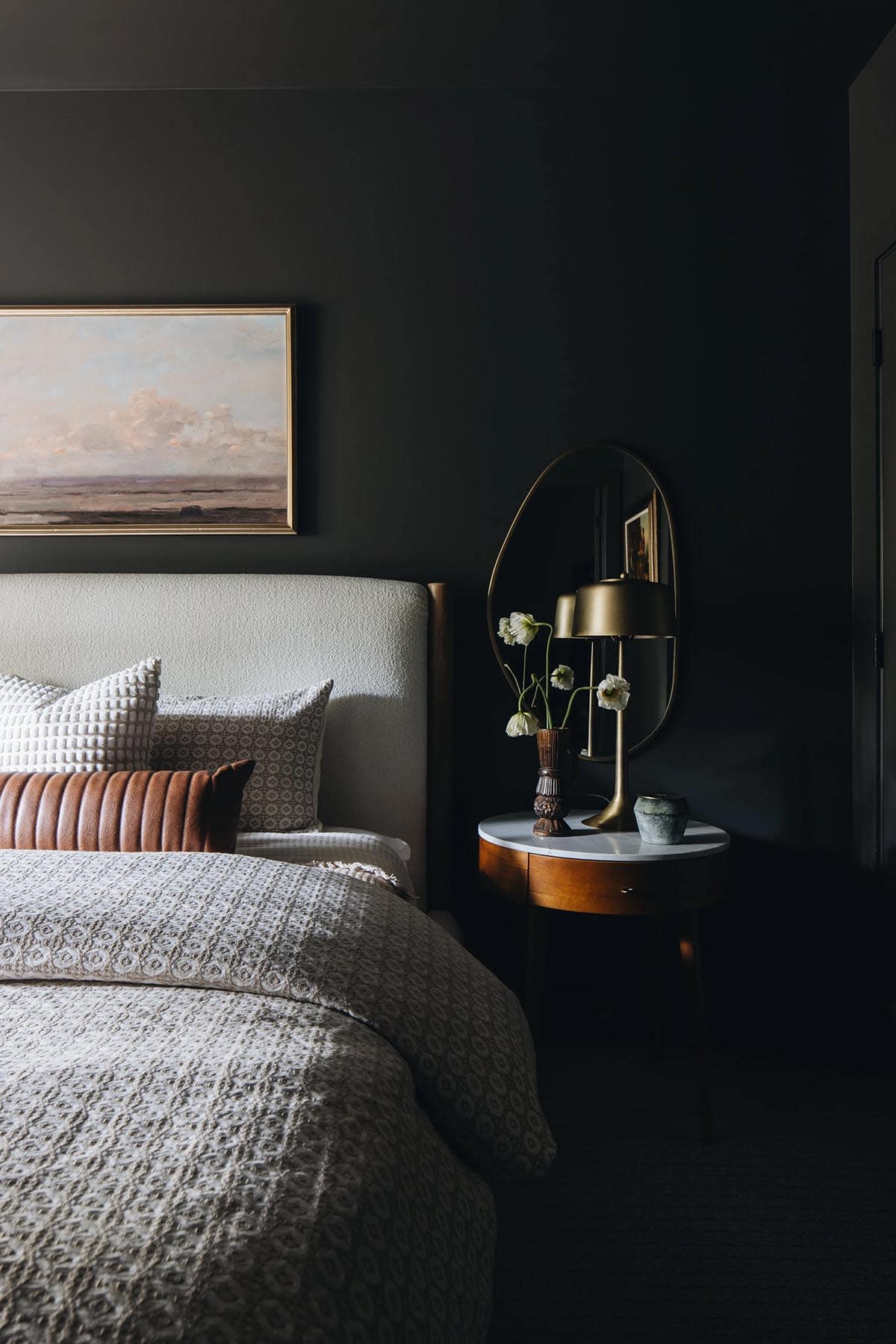 Dark bedroom nightstand vignette with a boucle wingback headboard, round walnut bedside table with marble top, brass table lamp, oval brass mirror, and white poppy arrangement against charcoal walls