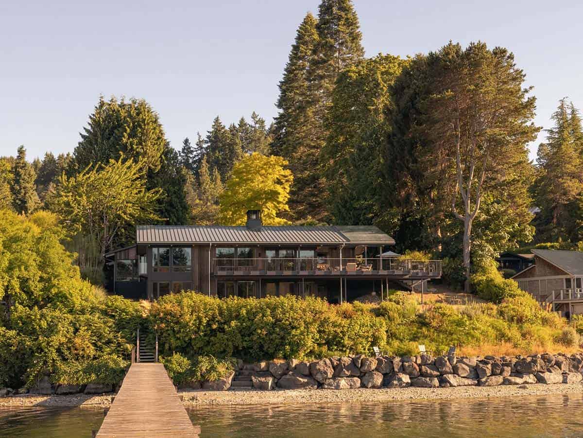 Waterfront home viewed from the dock, with dark wood siding, metal roof, and wraparound deck nestled among Pacific Northwest trees