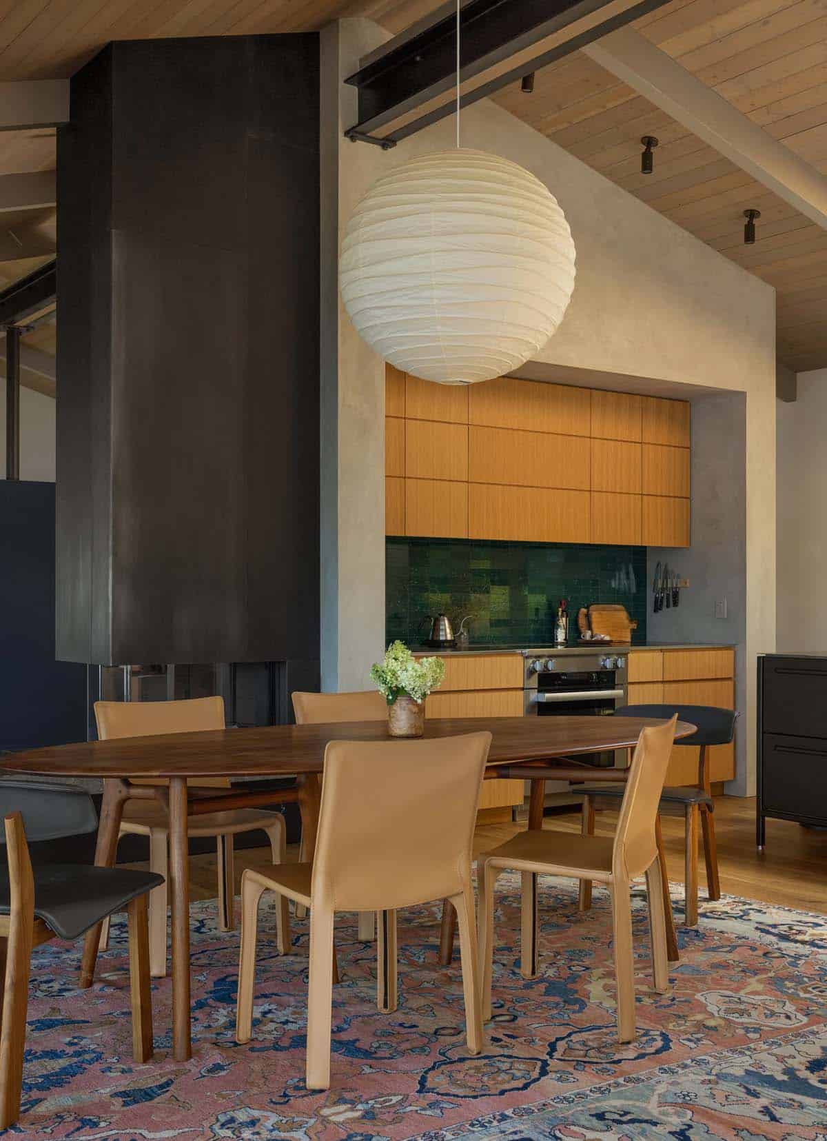 Open-plan dining area with walnut oval table, tan leather chairs, globe pendant light, and oak kitchen cabinets with green tile backsplash