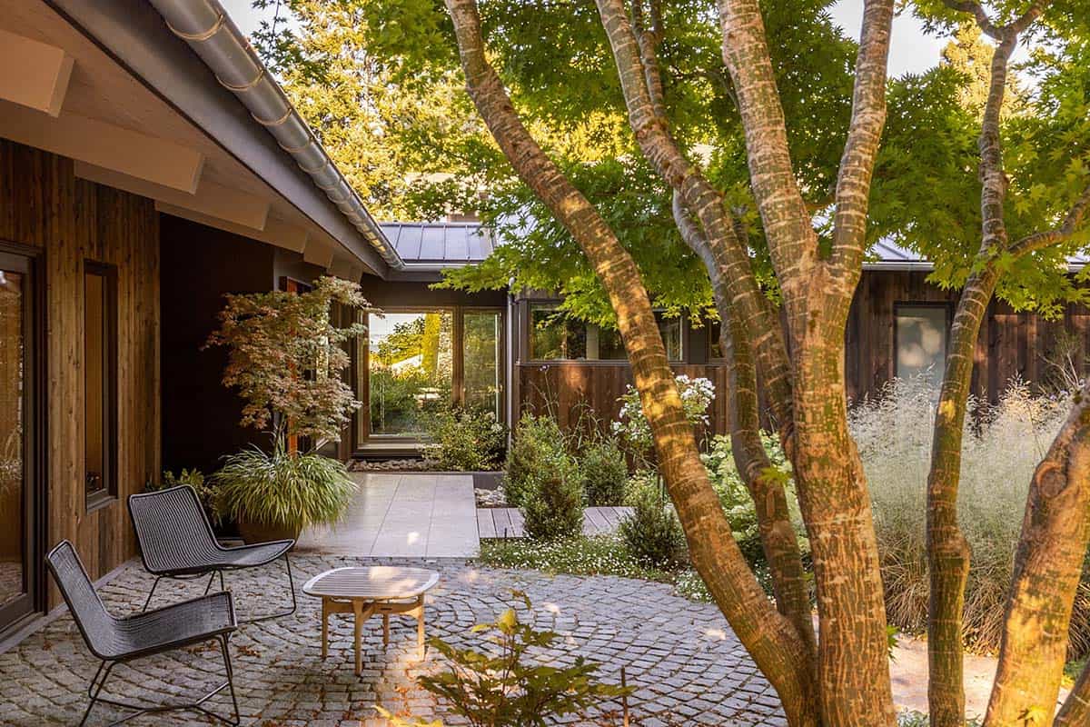 Private cobblestone courtyard with wire lounge chairs, Japanese maple trees, and glass connector between two wings of the home