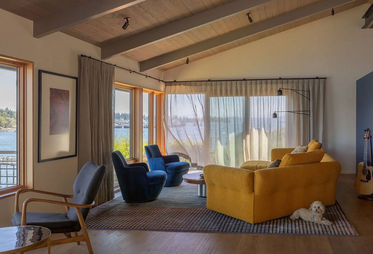 Living room with mustard yellow sofa, navy swivel chairs, sheer curtains, exposed wood beam ceiling, and water views beyond