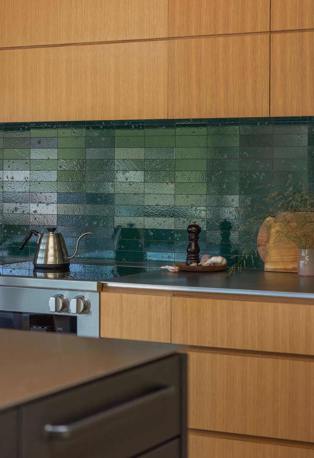 Close-up of kitchen with warm oak cabinetry, stainless steel countertop, and a richly textured green zellige tile backsplash