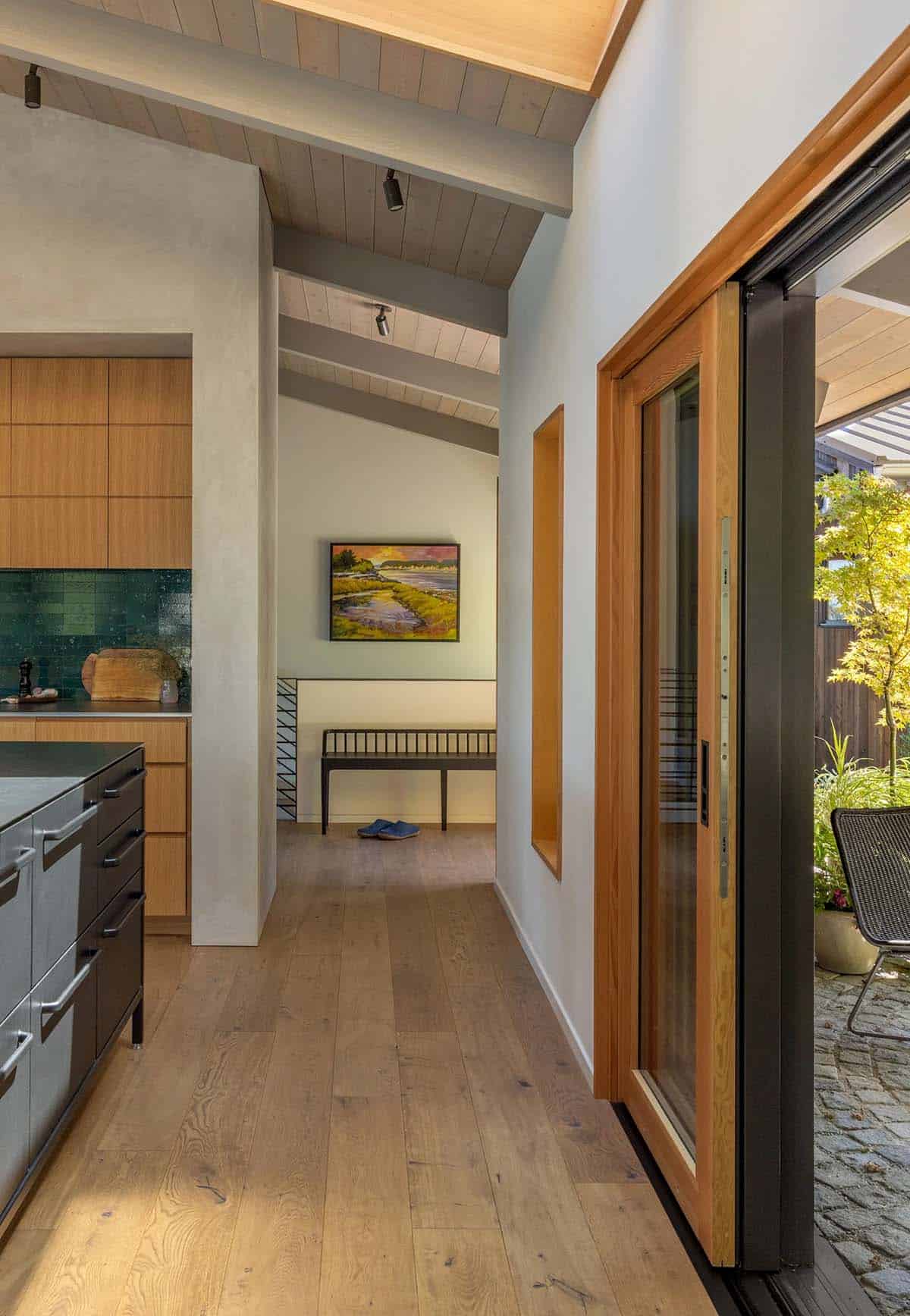 Interior hallway with wide-plank oak floors, exposed ceiling beams, and a sliding glass door opening to a cobblestone garden courtyard