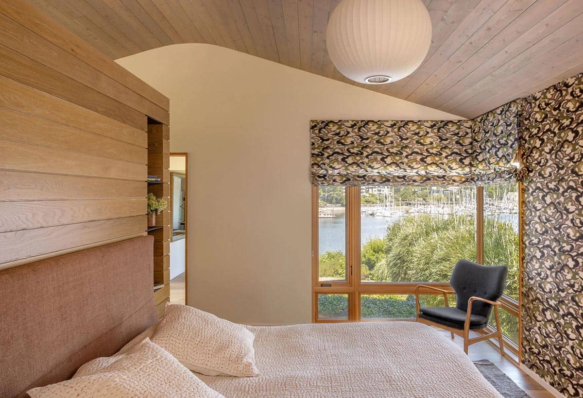 Primary bedroom with water views, patterned Roman shades and wallpaper, pink upholstered headboard, and globe pendant light