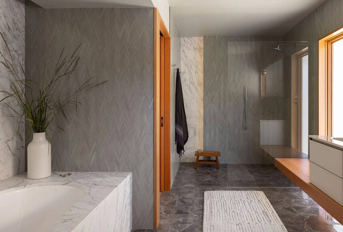 Primary bathroom with gray chevron tile walls, marble soaking tub surround, warm wood vanity, and walk-in shower