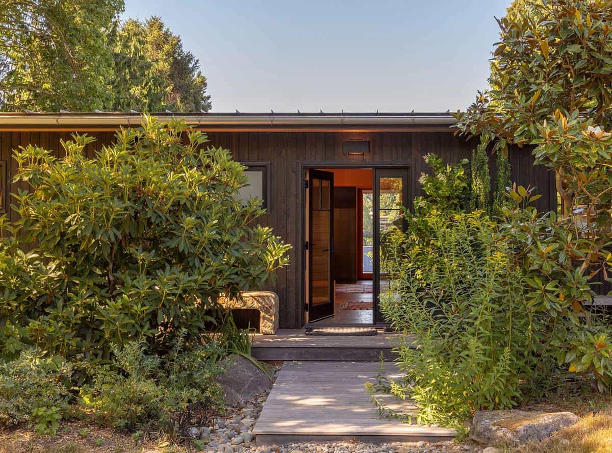 Guest cottage entrance with dark vertical wood siding, open pivot door, and overgrown native plantings along a boardwalk path