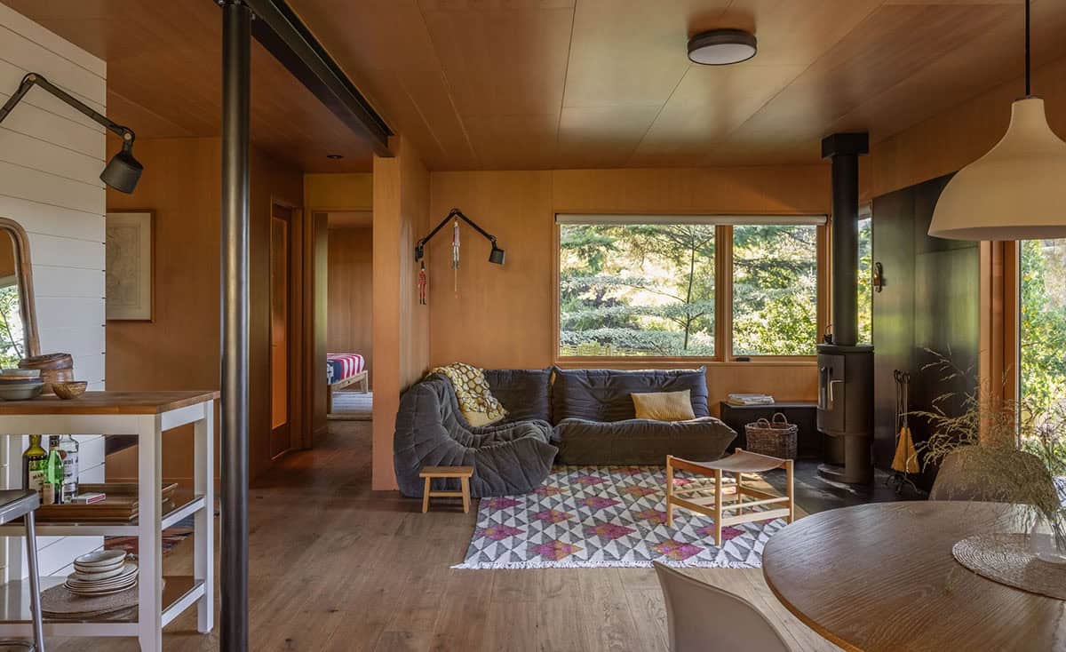 Guest cottage living area with terracotta walls, Togo-style sectional, wood stove, patterned rug, and garden views